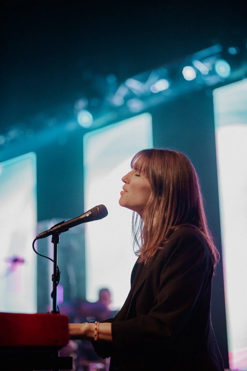 A woman with brown hair and a black blazer playing a red keyboard on stage, with her eyes closed and a microphone in front of her, during a performance or recording.