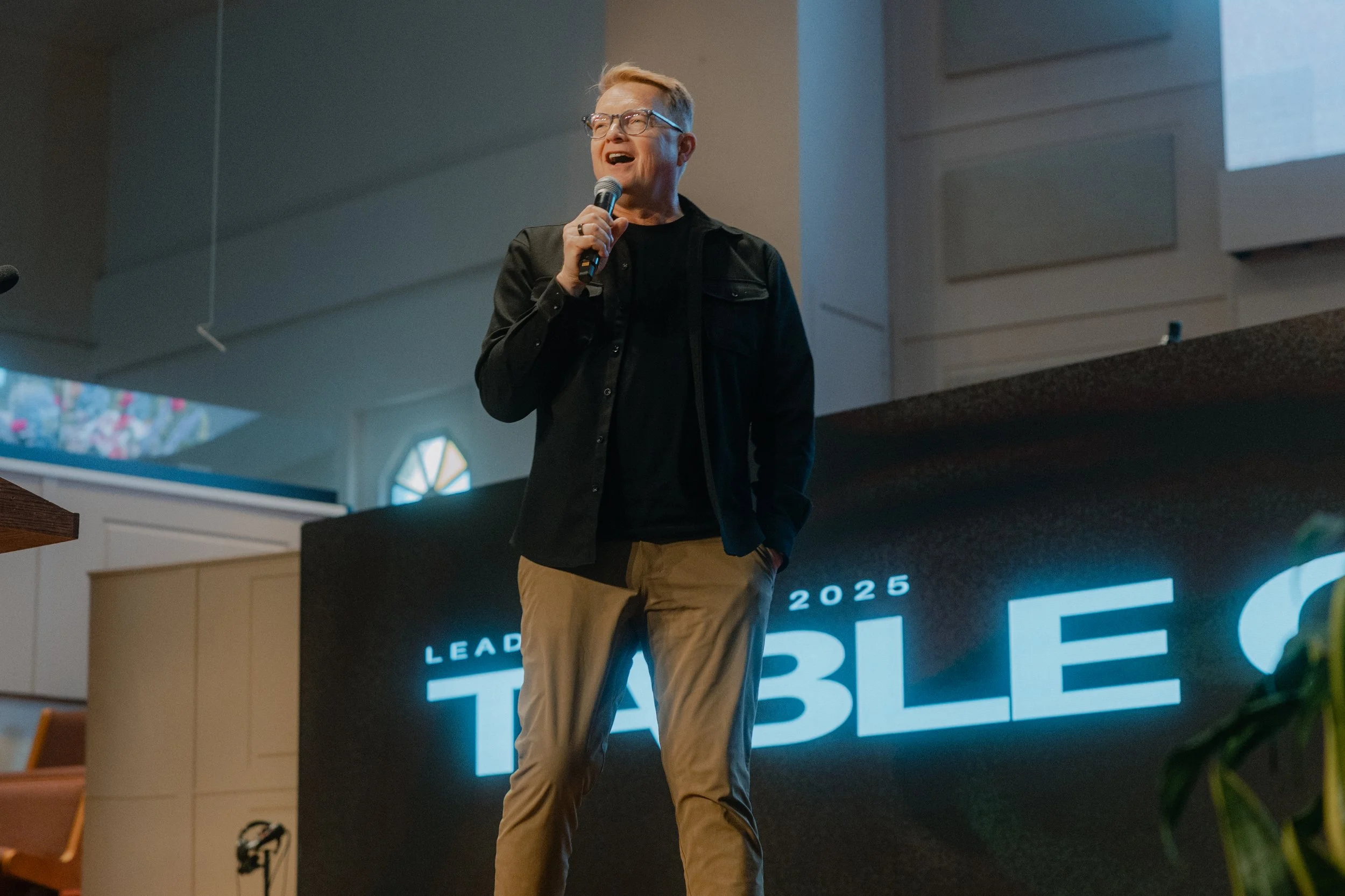 A man in glasses and black jacket speaking into a microphone on stage at a conference called 'LEAD TABLE 2025' with a large screen behind him showing the event name.