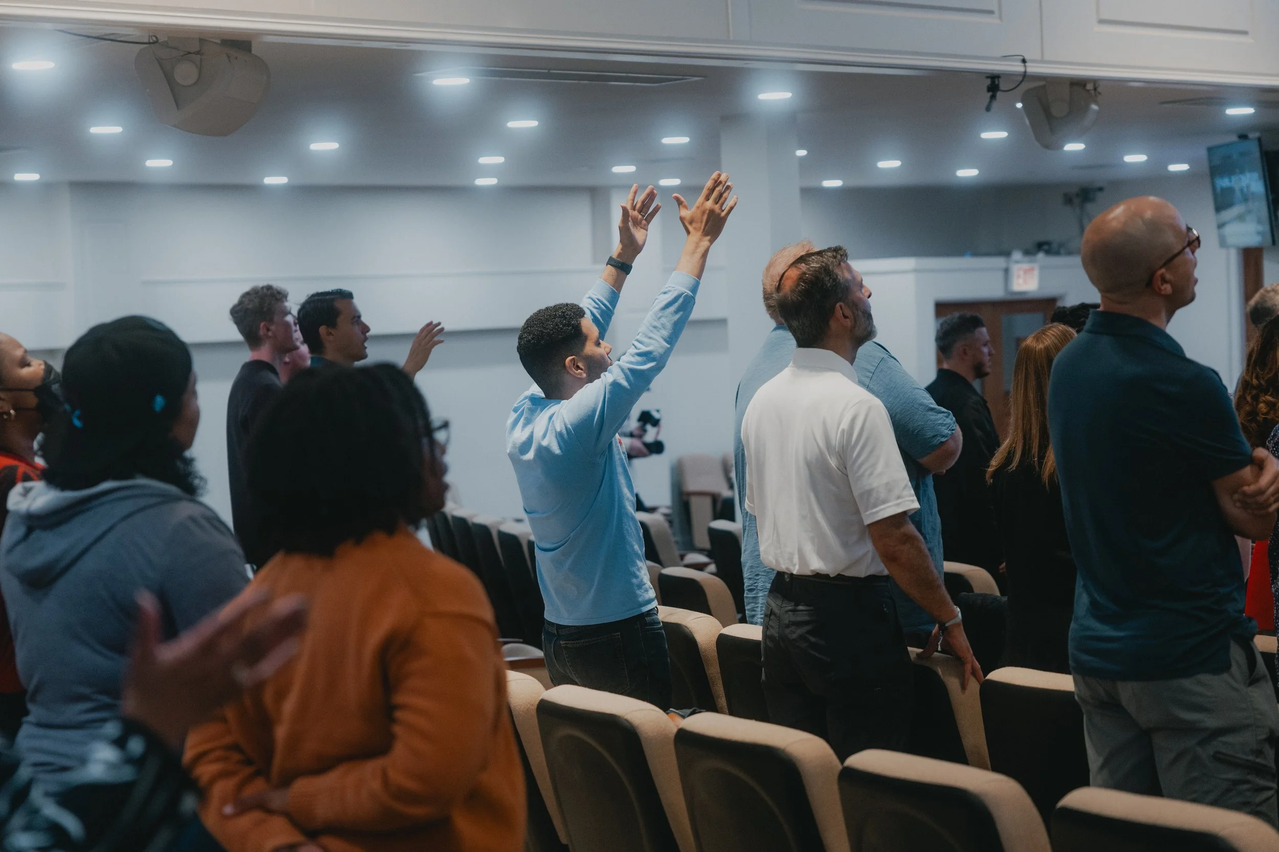 People attending a conference or event in a large room, some raising their hands, others standing attentively, with a screen visible in the background.