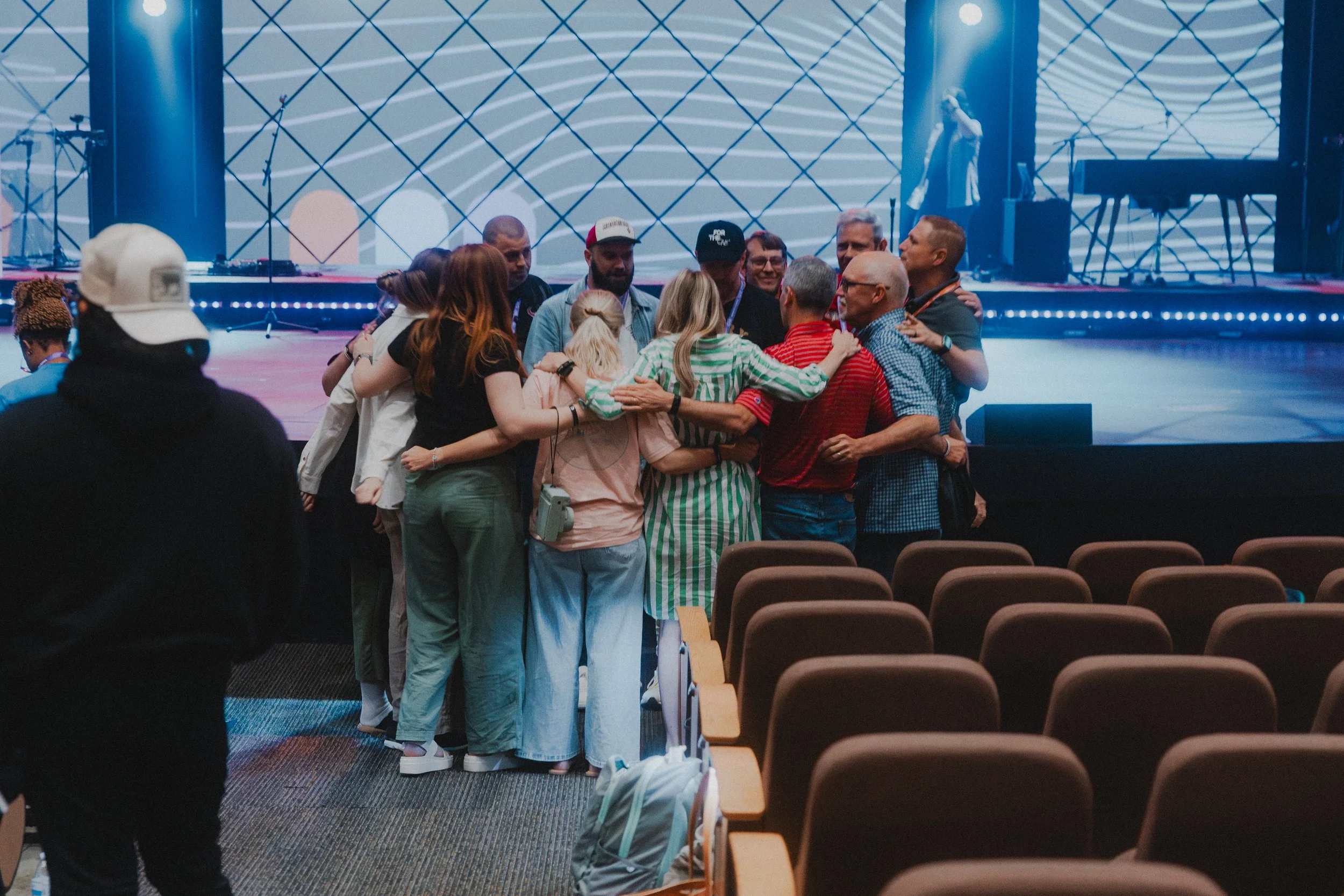 Group of people embracing and huddling together on a stage in an auditorium, with empty chairs in foreground and musical instruments on a stage in background.