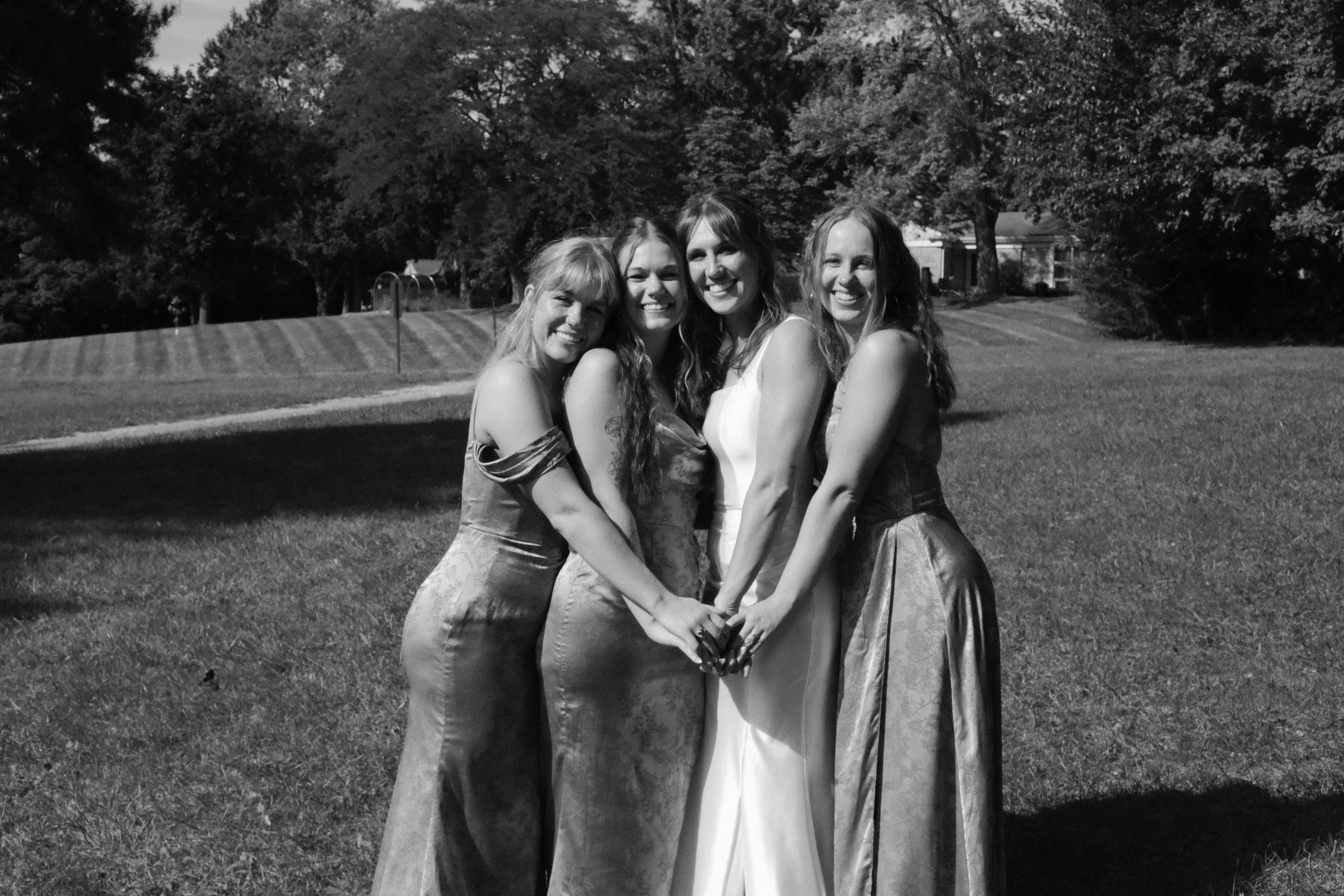 Four women in evening dresses standing closely together on a grassy field, holding hands, with trees and a house in the background.
