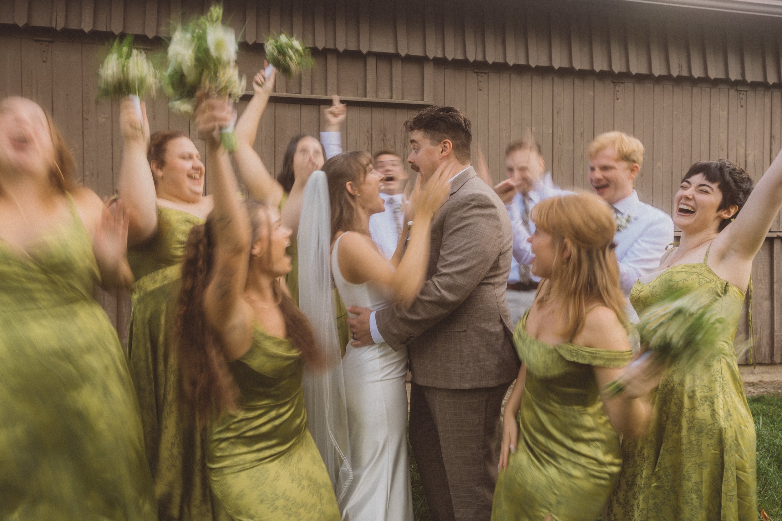 A wedding celebration outdoors with the bride and groom embracing in the center, surrounded by bridesmaids and groomsmen cheering and smiling, with bouquets raised in the air.