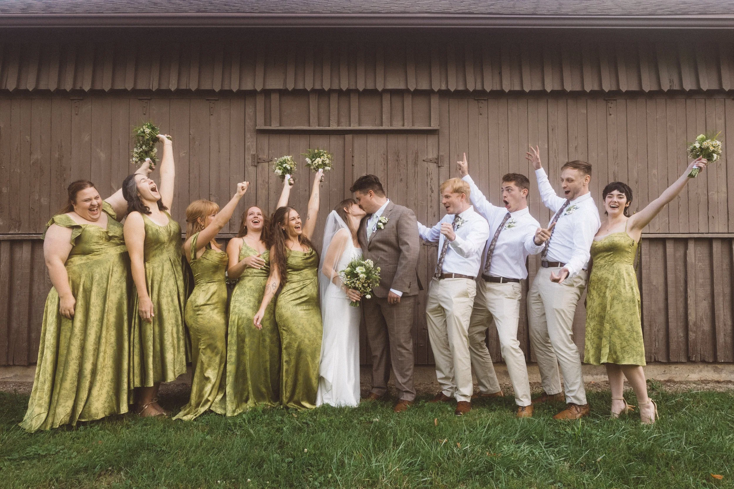 A wedding party with bridesmaids in green dresses and groomsmen in white shirts and beige pants, celebrating and cheering around a bride and groom who are kissing. The bridesmaids are holding bouquets and raising their hands in excitement, standing i