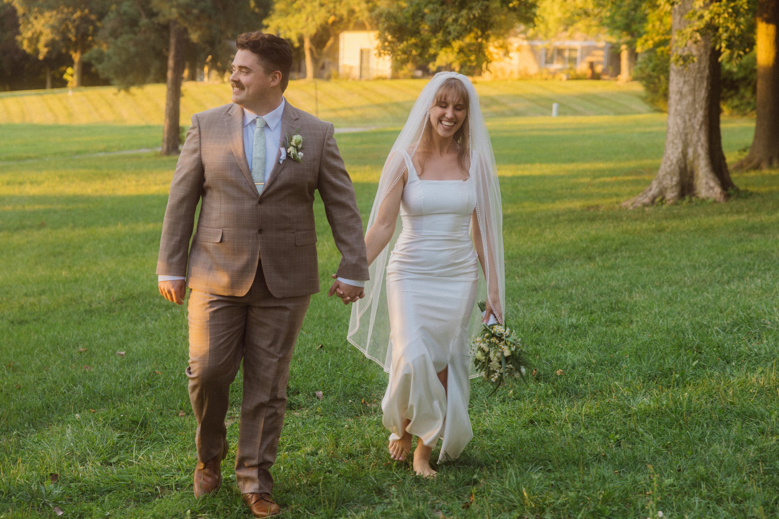 A newlywed couple walking hand-in-hand on a grassy field during sunset. The man is wearing a brown plaid suit with a light blue tie and boutonniere. The woman is in a white wedding gown with a veil, holding a small bouquet of flowers, smiling happily