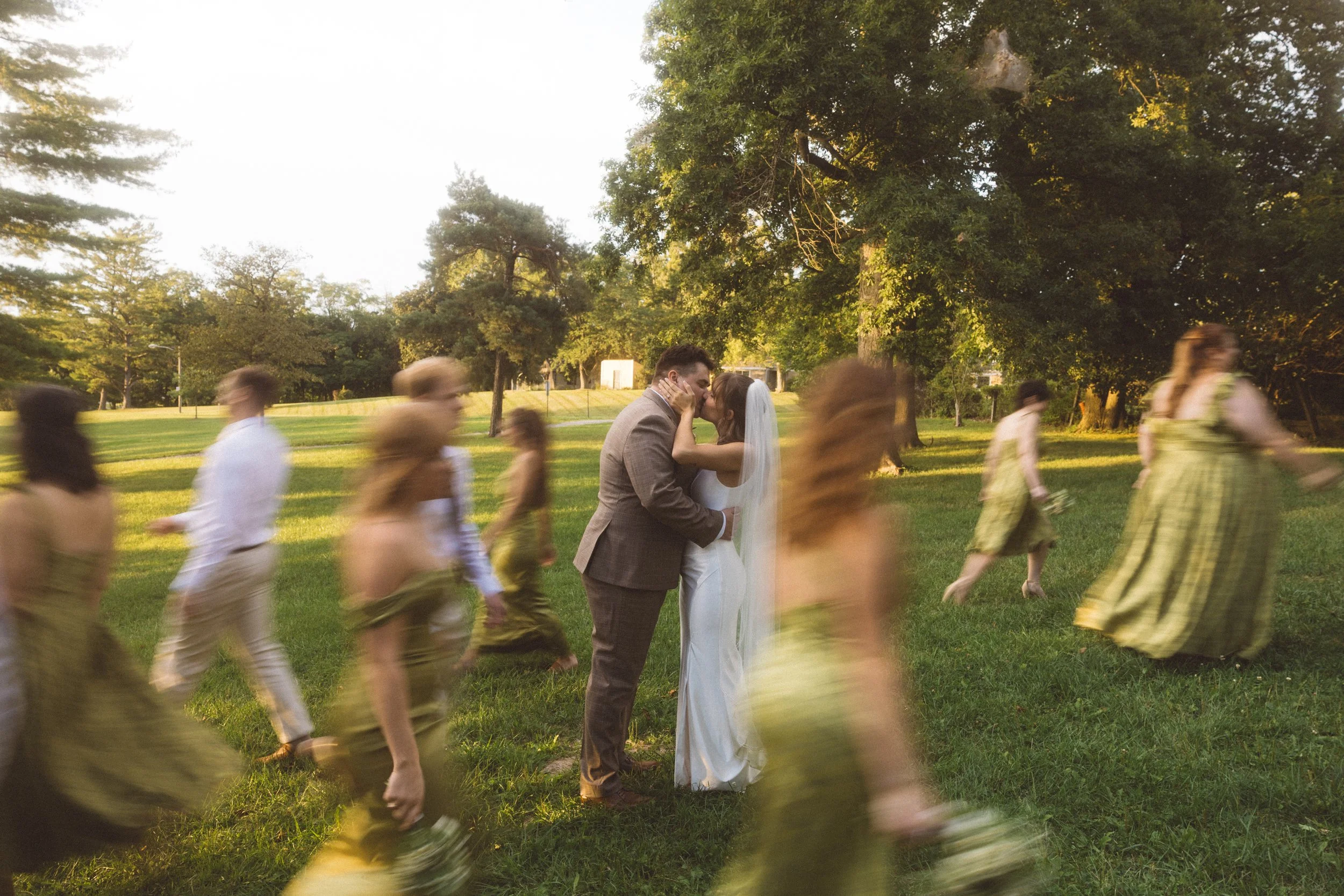 A bride and groom sharing a kiss in the center, surrounded by blurred figures of people in motion, set in a park with green grass and trees, during sunset.