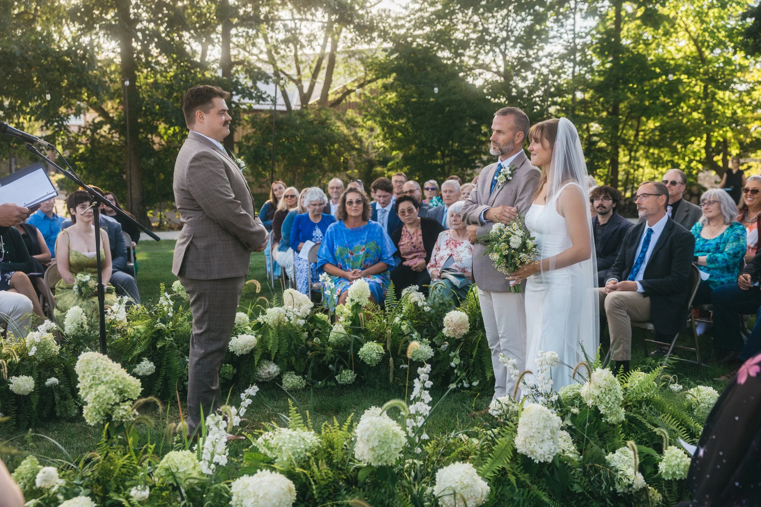 A wedding ceremony outdoors with the bride and groom standing in front of an officiant. The bride is holding a bouquet, and the groom is facing her. Guests are seated on either side, watching the ceremony amidst greenery and flowers during the daytim