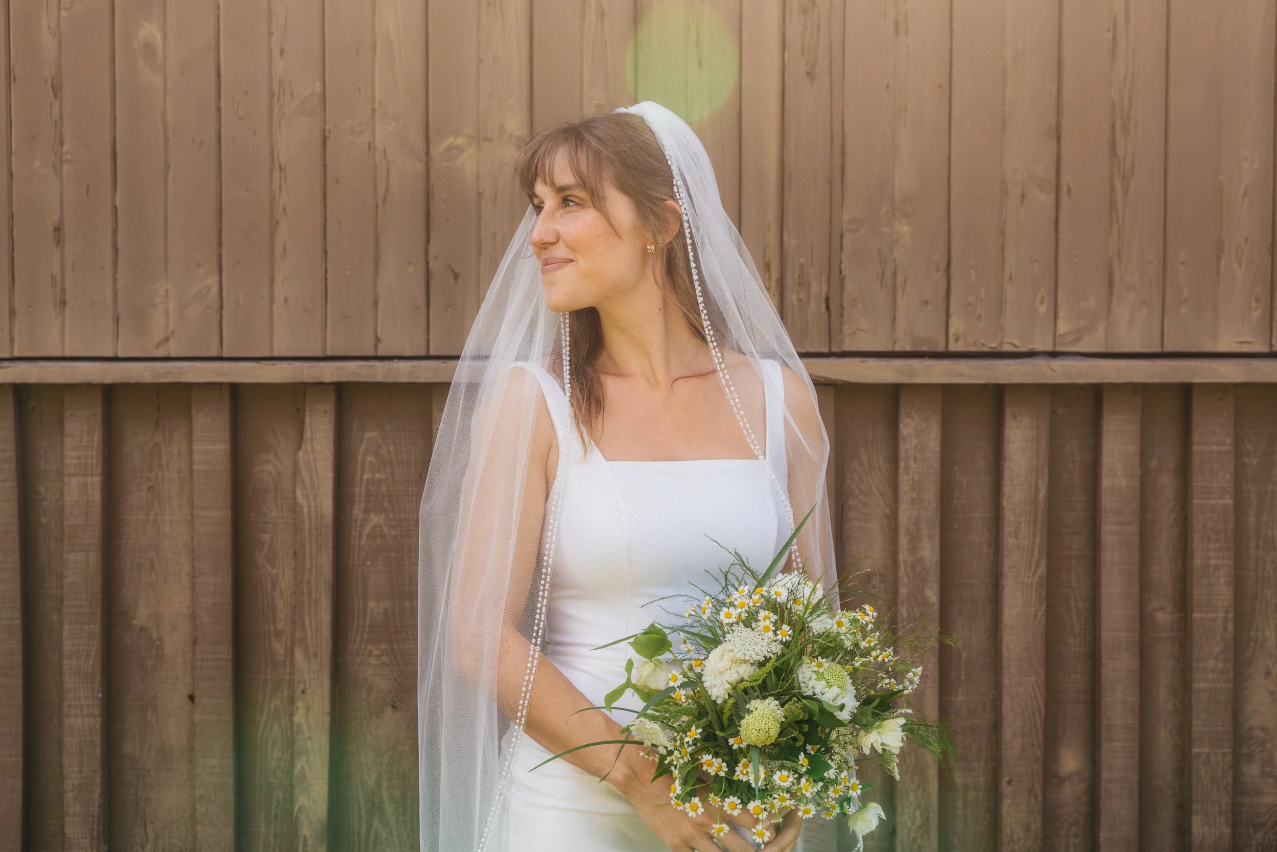 A woman in a white wedding dress holding a bouquet of flowers, standing outdoors in front of a wooden fence