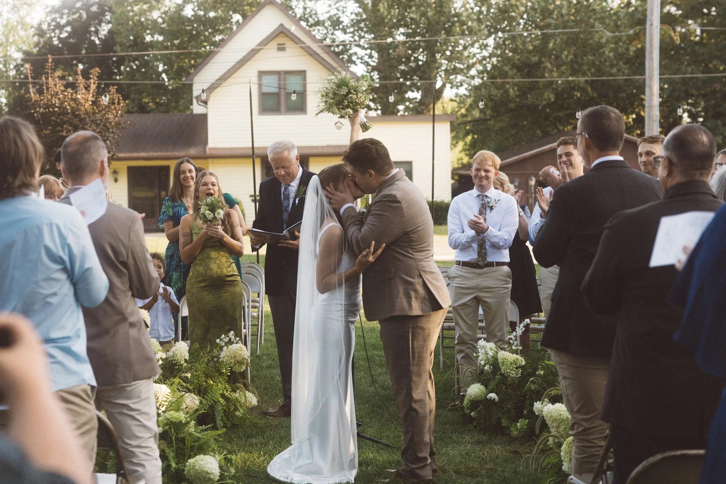 A wedding ceremony outdoors with a bride and groom sharing a kiss, surrounded by friends and family clapping and celebrating. The bride is holding a bouquet up in the air and the groom is gently holding her face as they kiss. The scene takes place on
