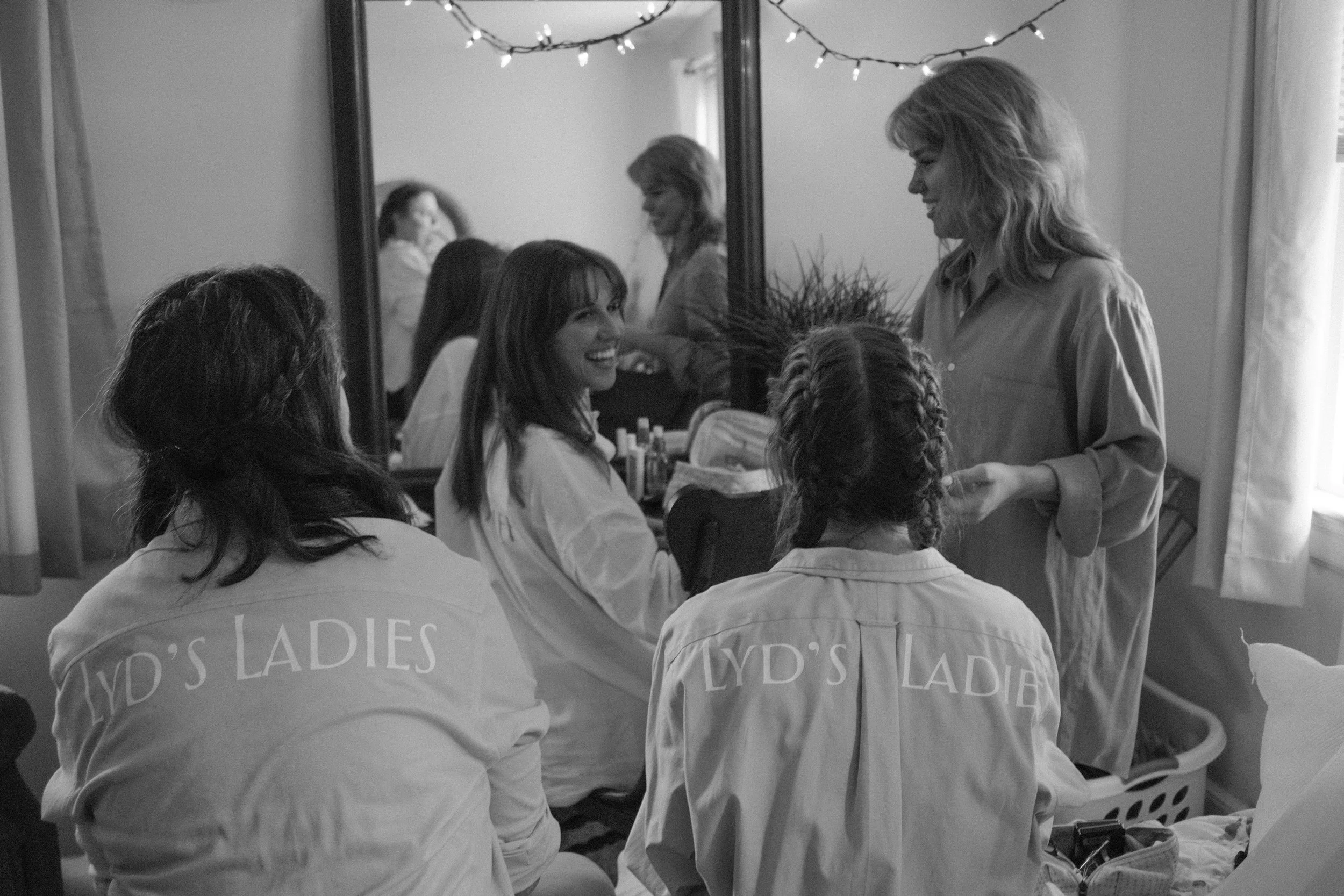 A group of women, with two wearing shirts labeled 'LYD'S LADIES,' are sitting in front of a mirror, smiling and talking. One woman is standing and holding some items, possibly preparing for a celebration or event. The room has fairy lights hanging ac