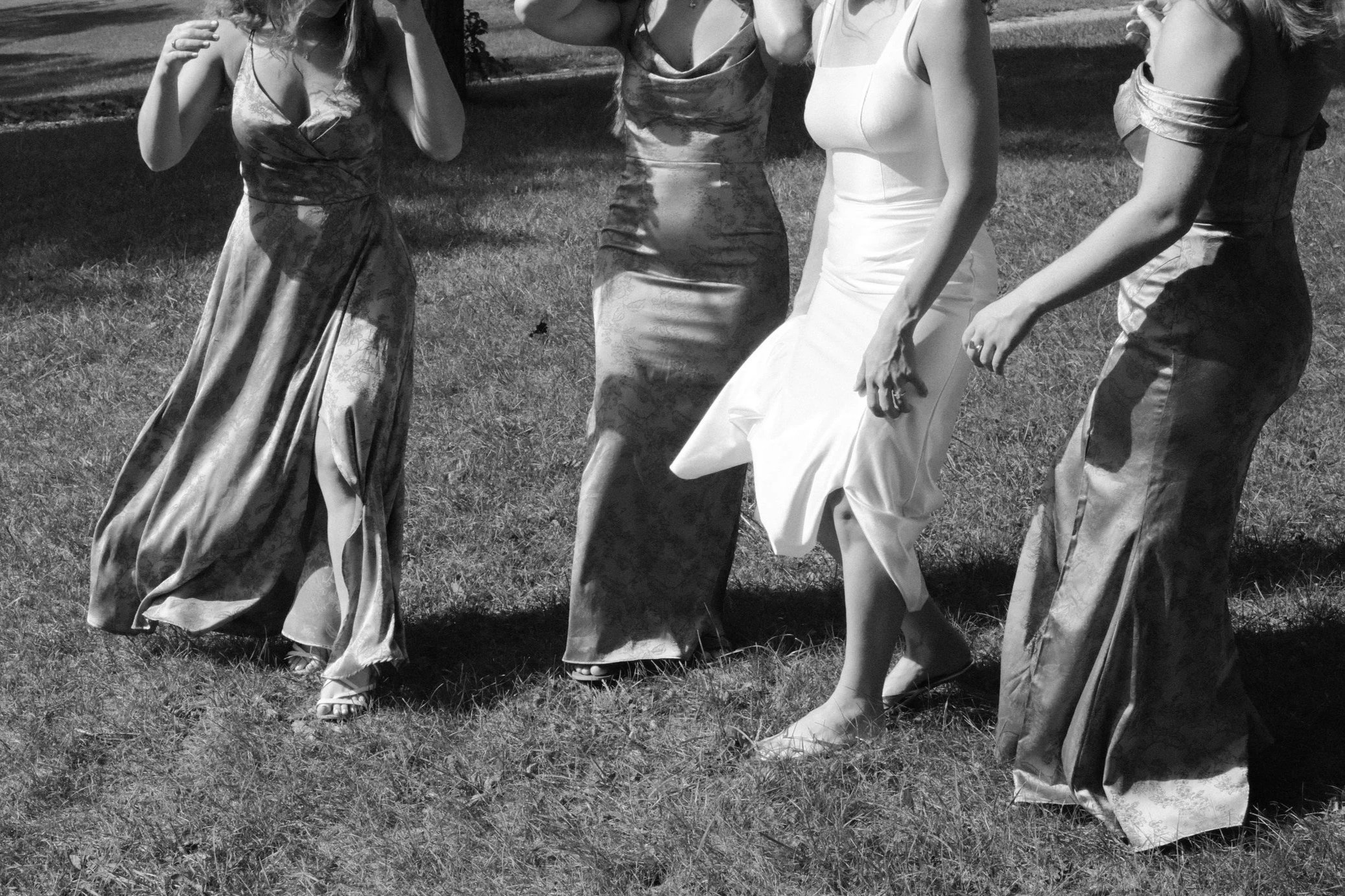 Four women in elegant dresses walking on a grassy area outdoors.