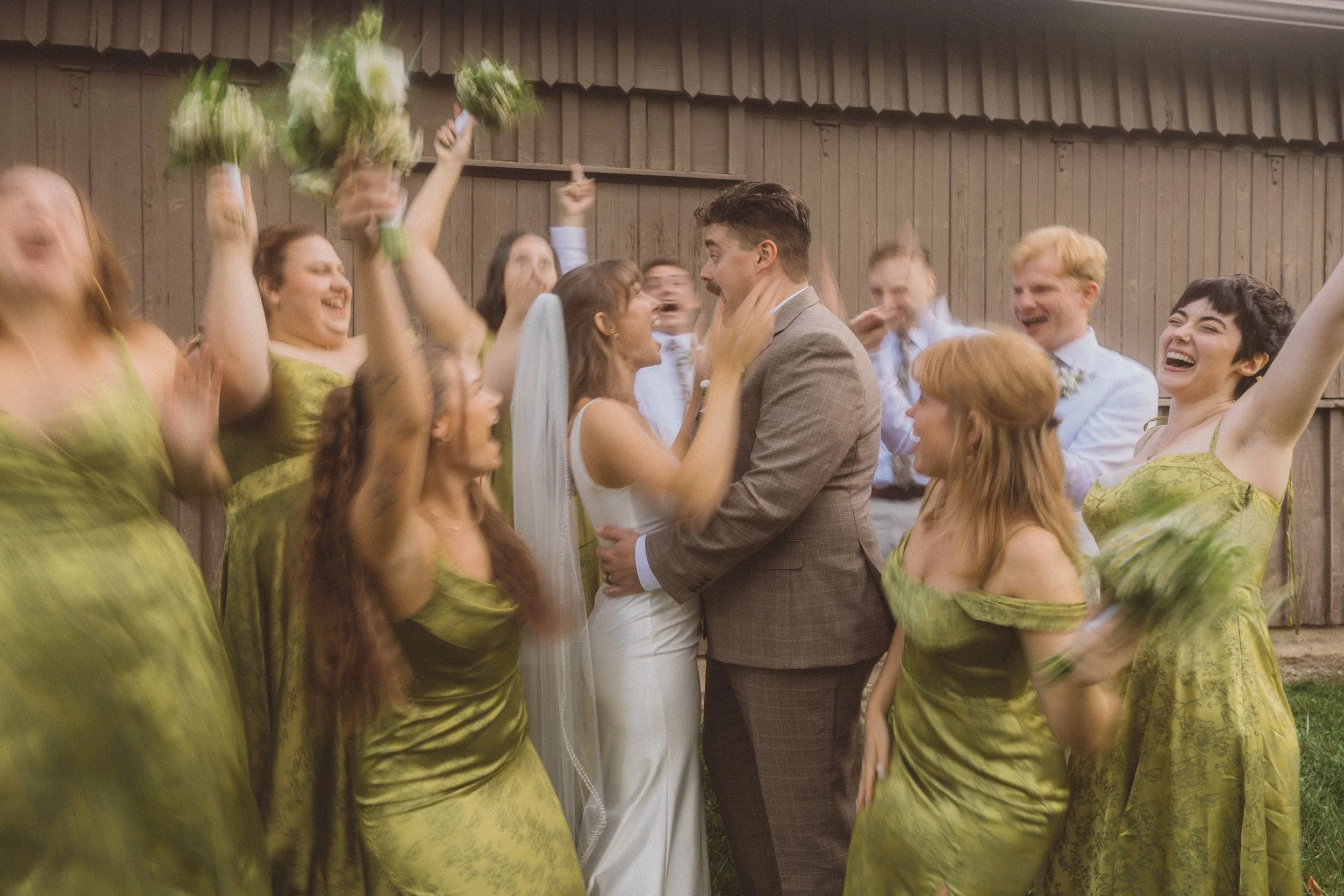 A bride and groom are holding each other and smiling, surrounded by laughing friends in green dresses and white shirts, celebrating outdoors near a wooden wall.
