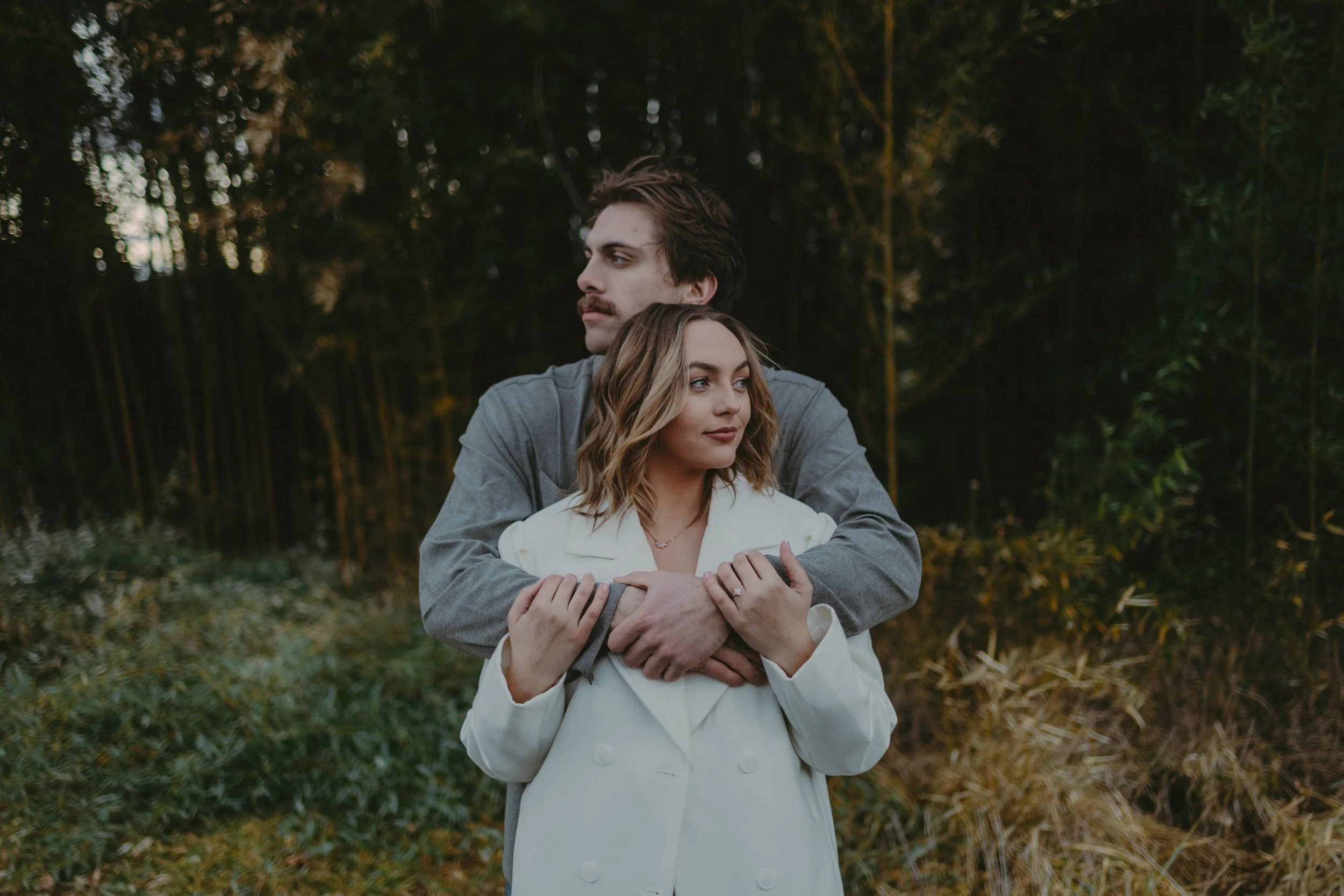 A man and woman standing closely together outdoors, with the man hugging the woman from behind. They are in a wooded area during the evening.