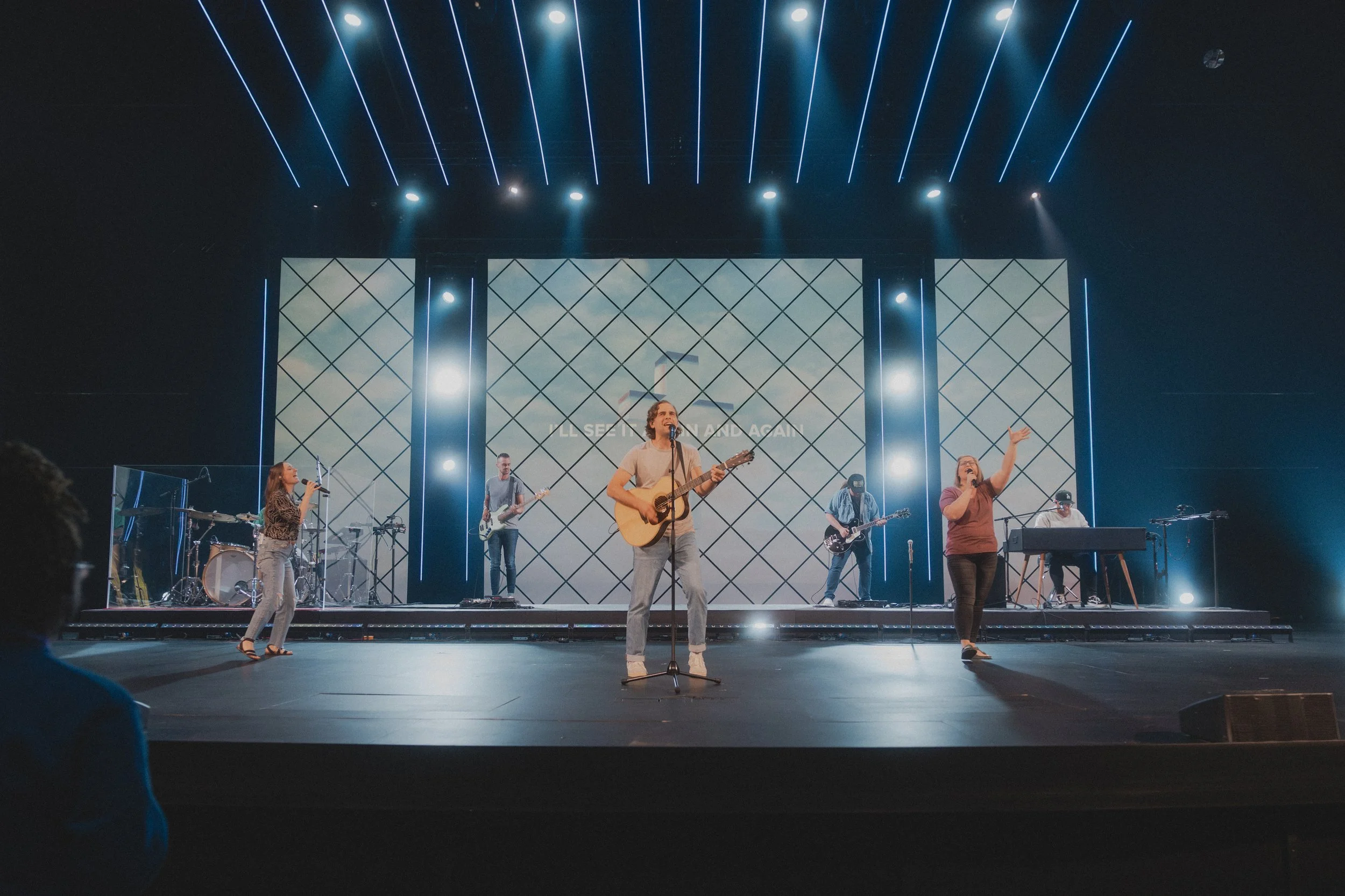 A band performing on stage with five members, including a woman singing into a microphone, a man playing an acoustic guitar, a woman singing with her arm raised, a man playing bass, and a man at keyboard, with large screens and stage lighting in the background.