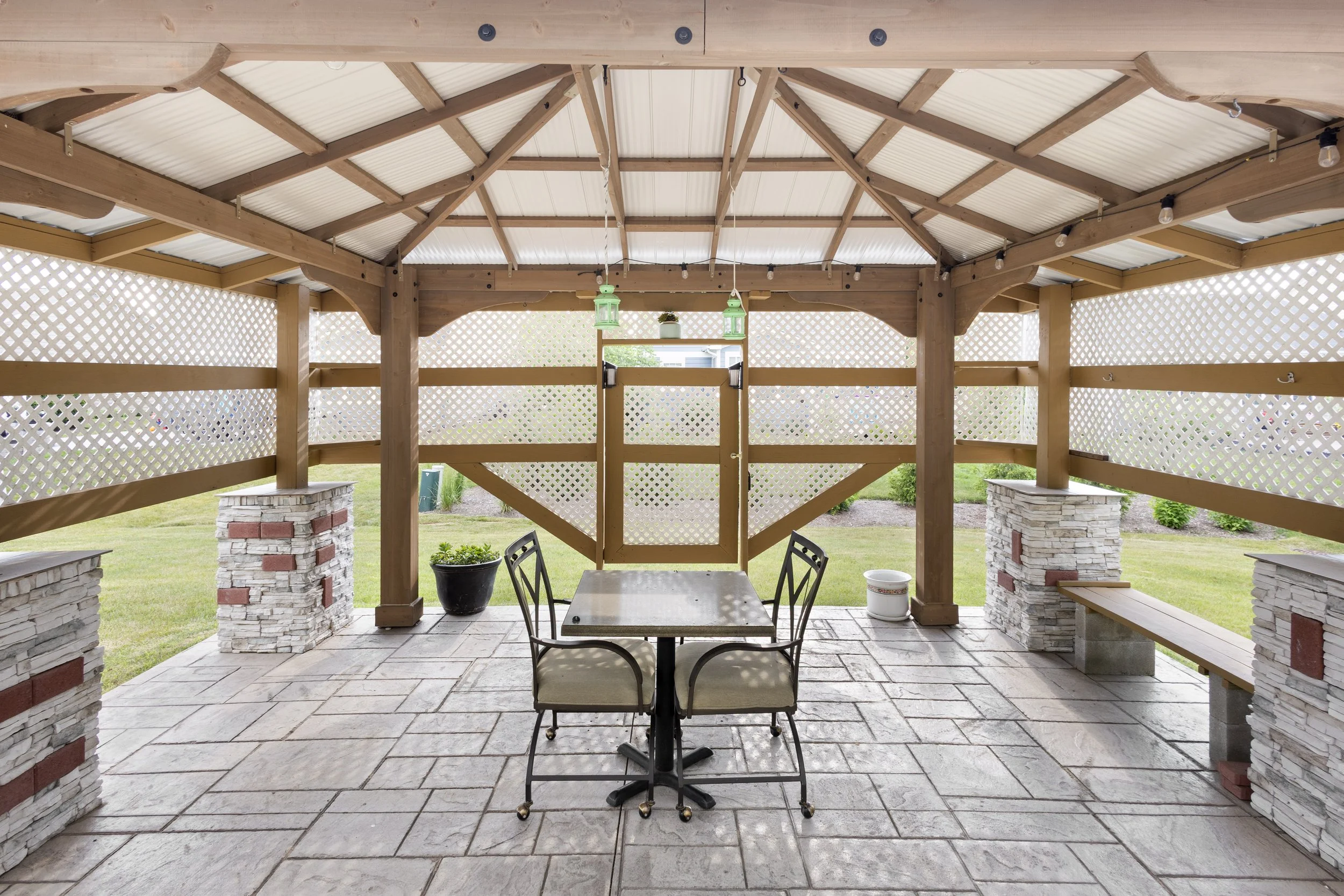 Covered outdoor patio with stone paving, wooden roof, lattice walls, a table with two chairs, and decorative lanterns hanging from the ceiling.
