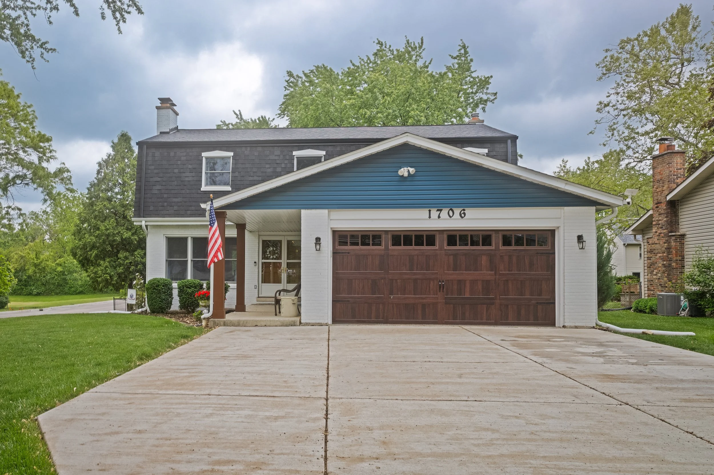 Front view of a two-story house with white and blue exterior, brown garage door, concrete driveway, and American flag on the porch, surrounded by green lawn and trees.