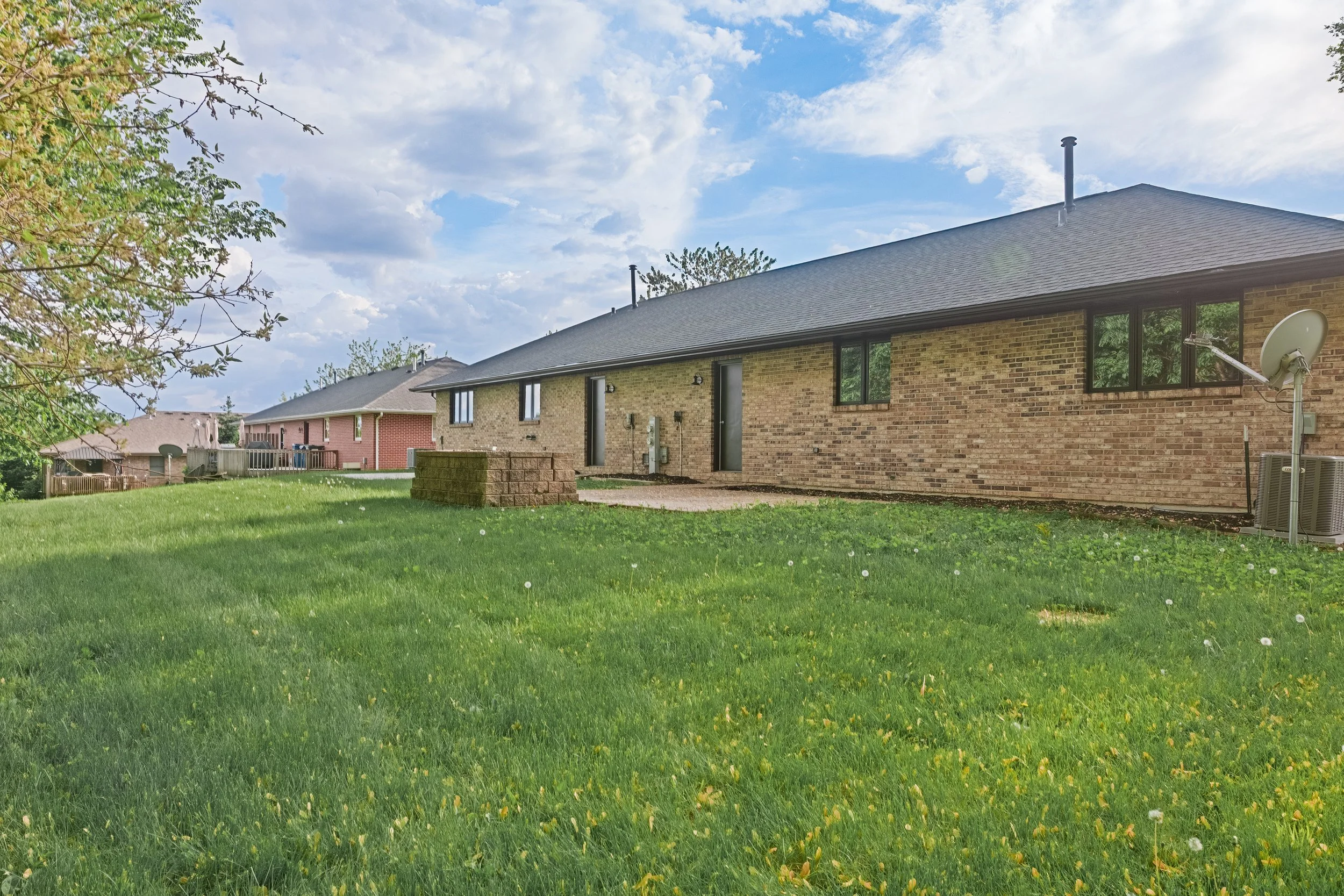 Backyard with green grass, a small brick platform, and a brick house with black window frames, a satellite dish, and an air conditioning unit. A tree with budding leaves is on the left, and the sky is partly cloudy.