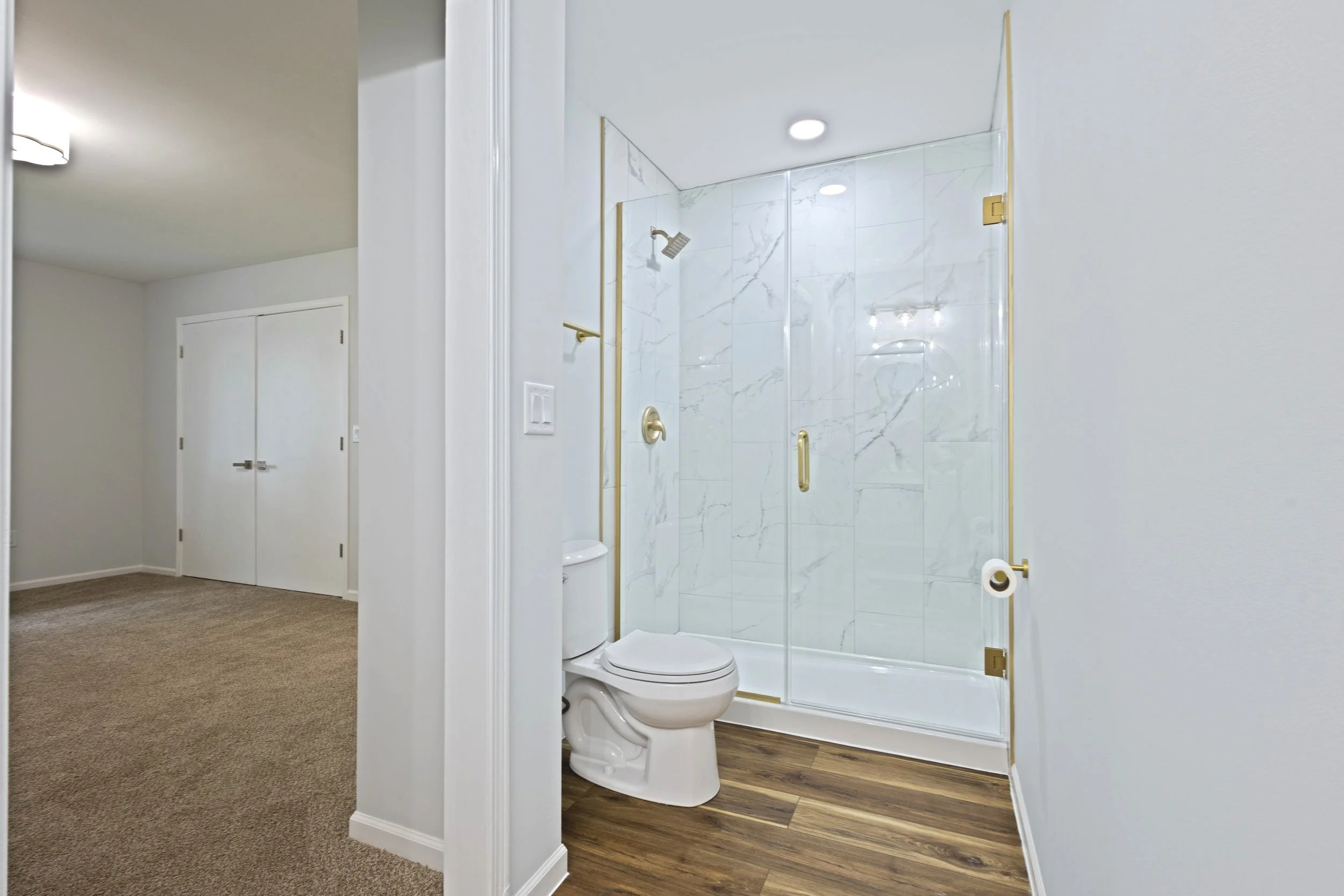 Bathroom with a glass enclosed shower, a white toilet, and wood flooring, adjacent to a carpeted room with white doors.
