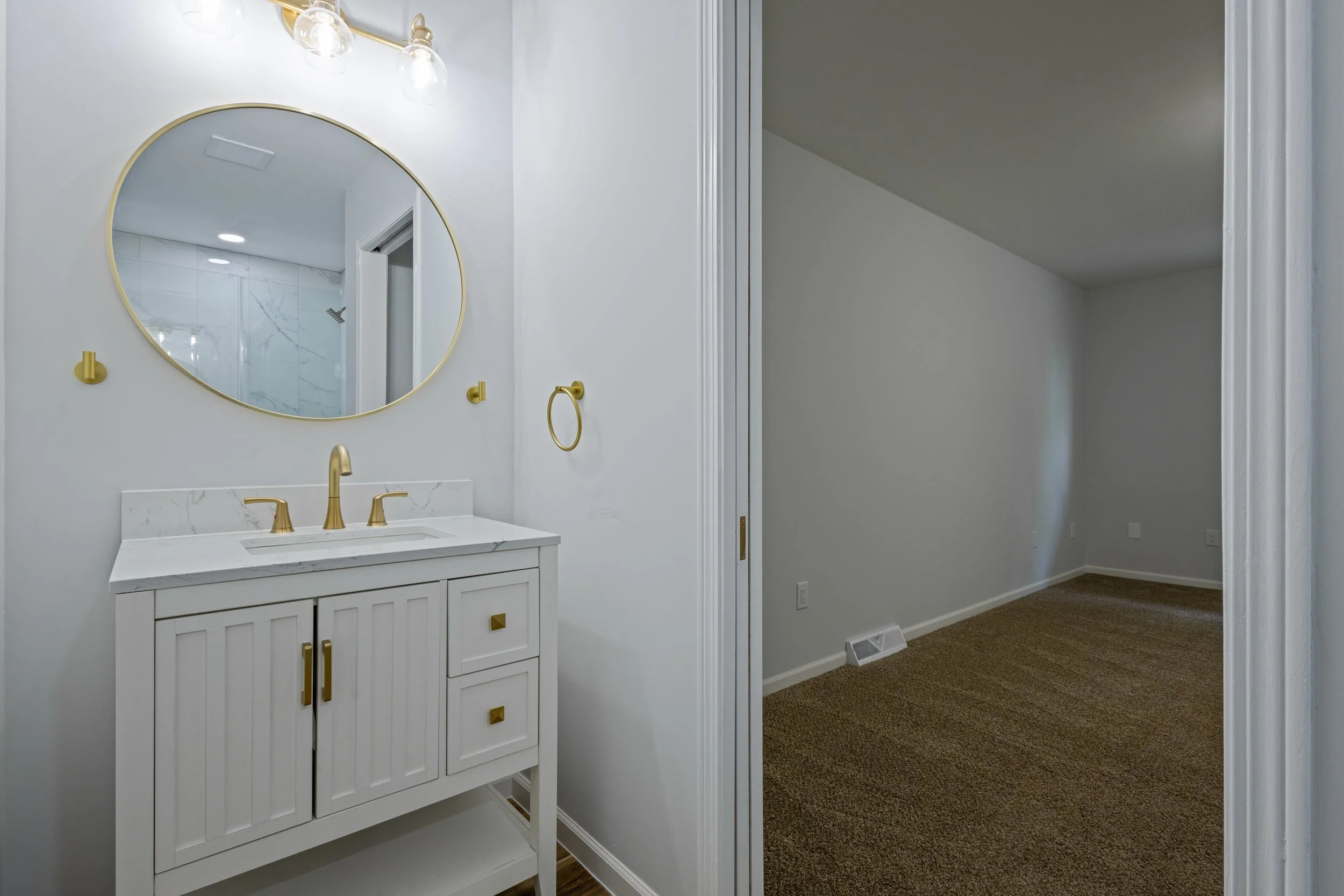 Bathroom vanity with a large round mirror, gold fixtures, and a white cabinet. An adjacent room with brown carpet is visible.
