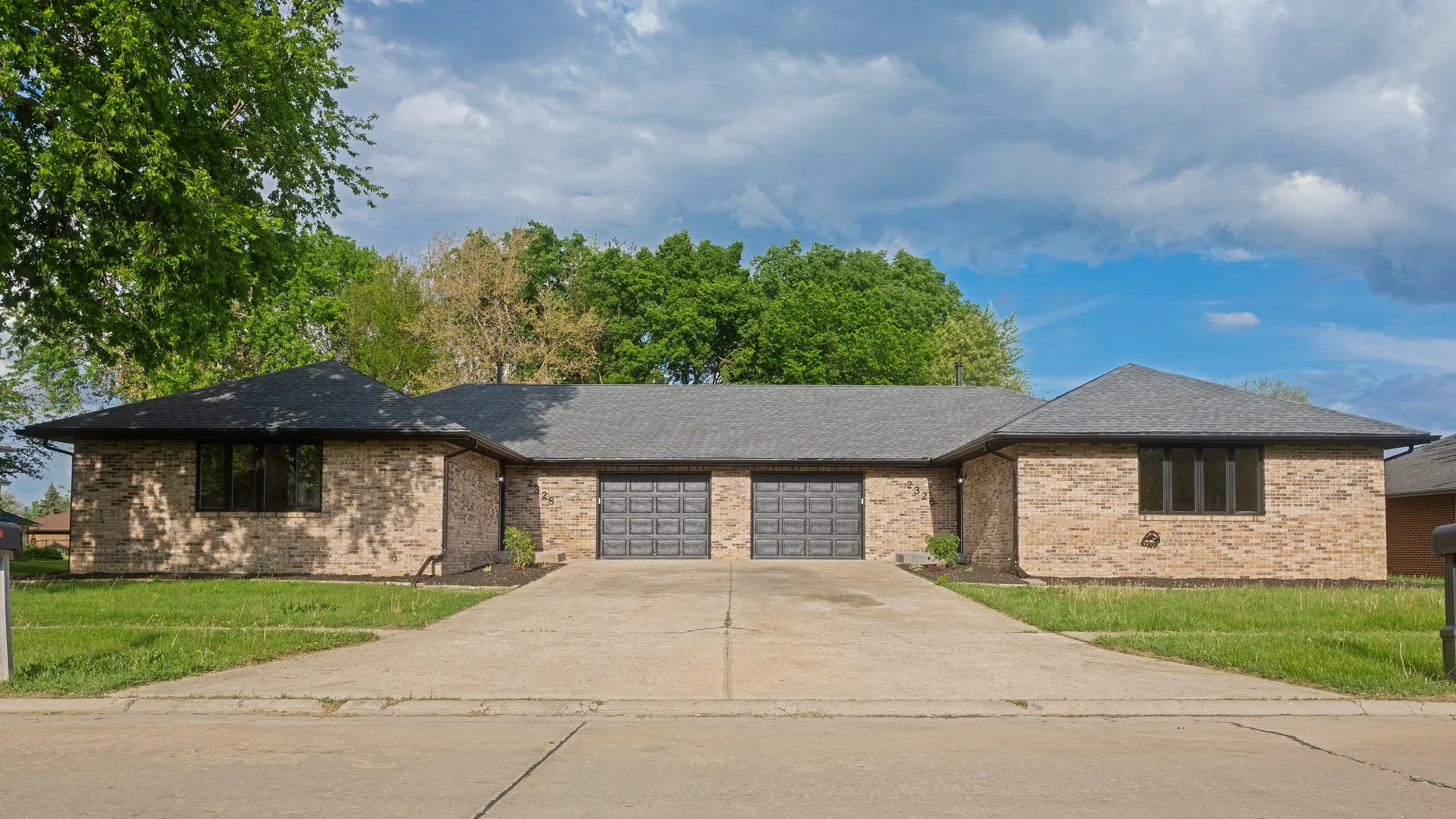 A single-story brick house with two garage doors, a driveway, green lawn, large trees in the background, and a cloudy sky.