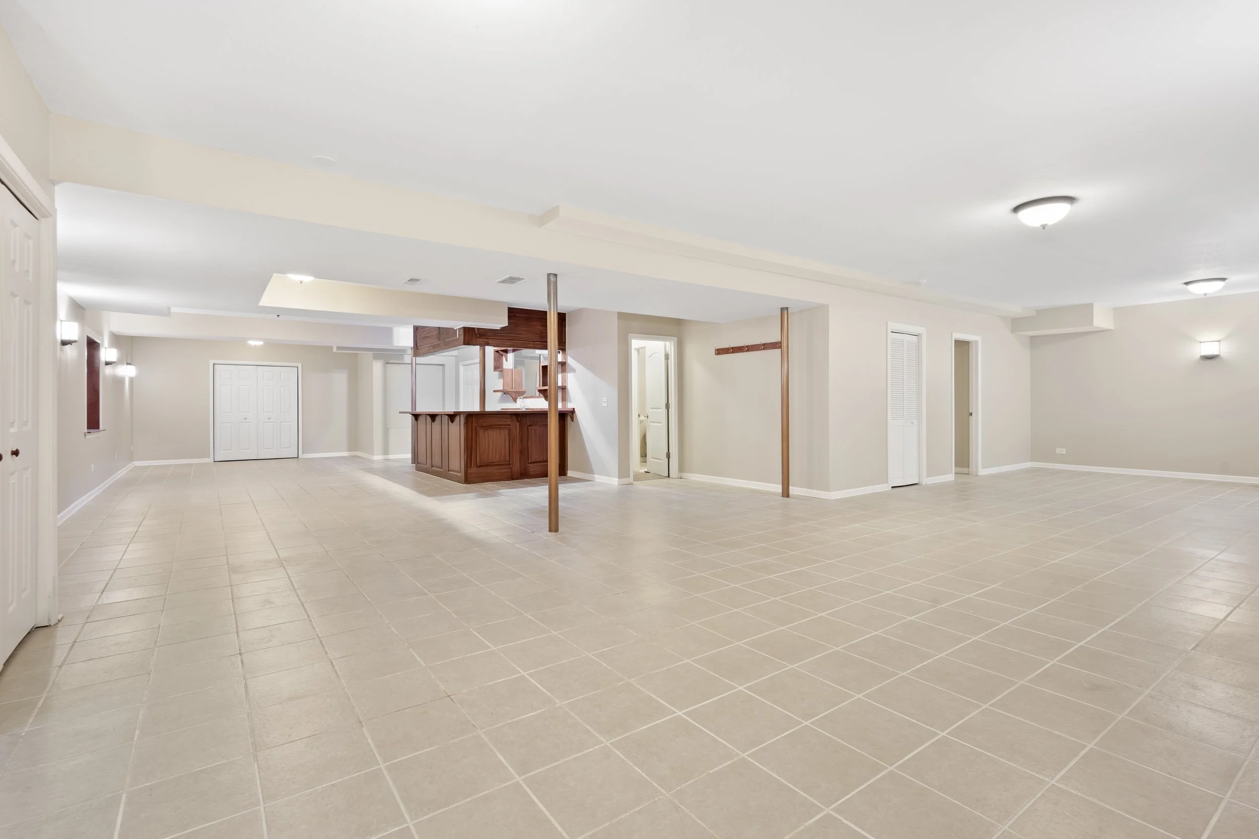 Empty spacious basement with beige tiled floors, cream walls, and multiple ceiling lights, featuring a wooden bar area and doors leading to other rooms.