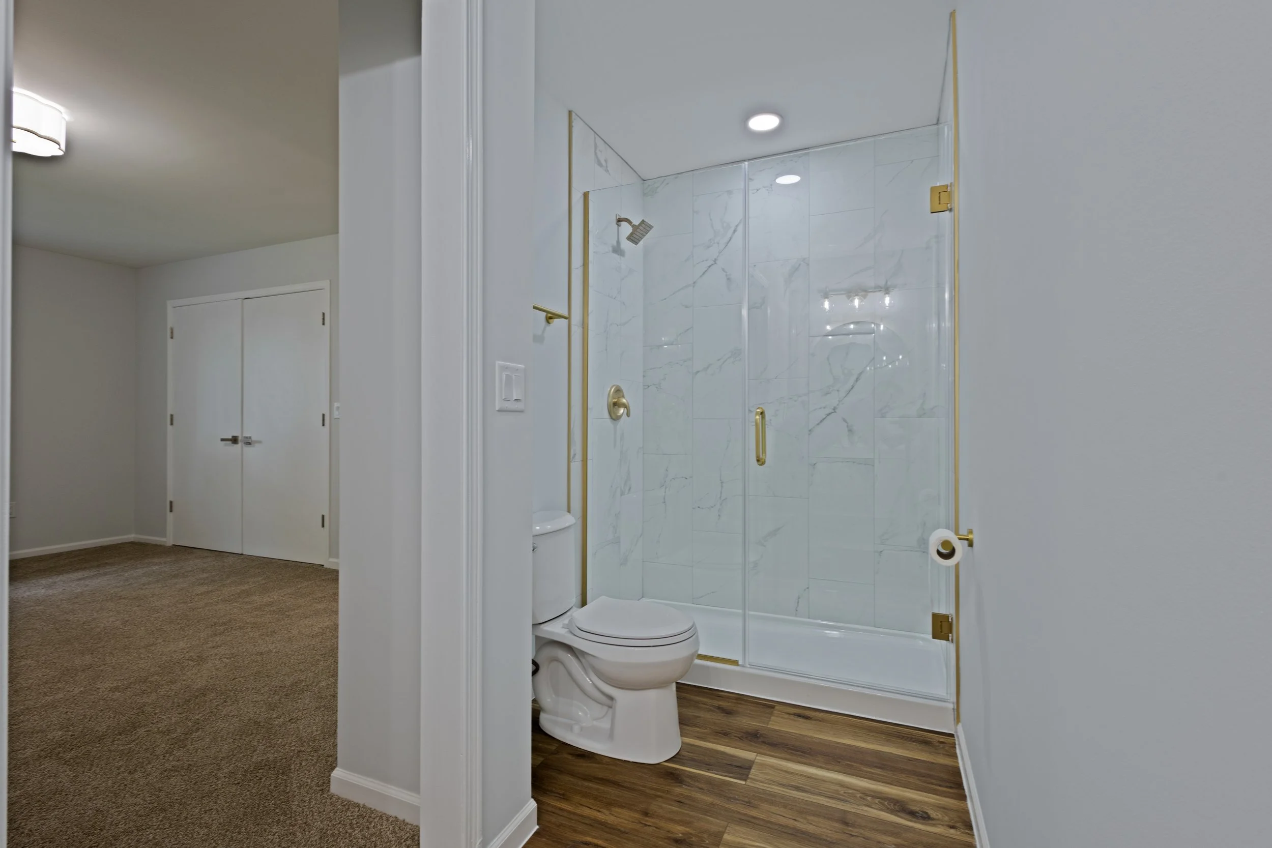 Bathroom with a glass shower door with gold accents, a white toilet, and wood-look vinyl flooring, adjacent to a carpeted bedroom with white walls and closet doors.