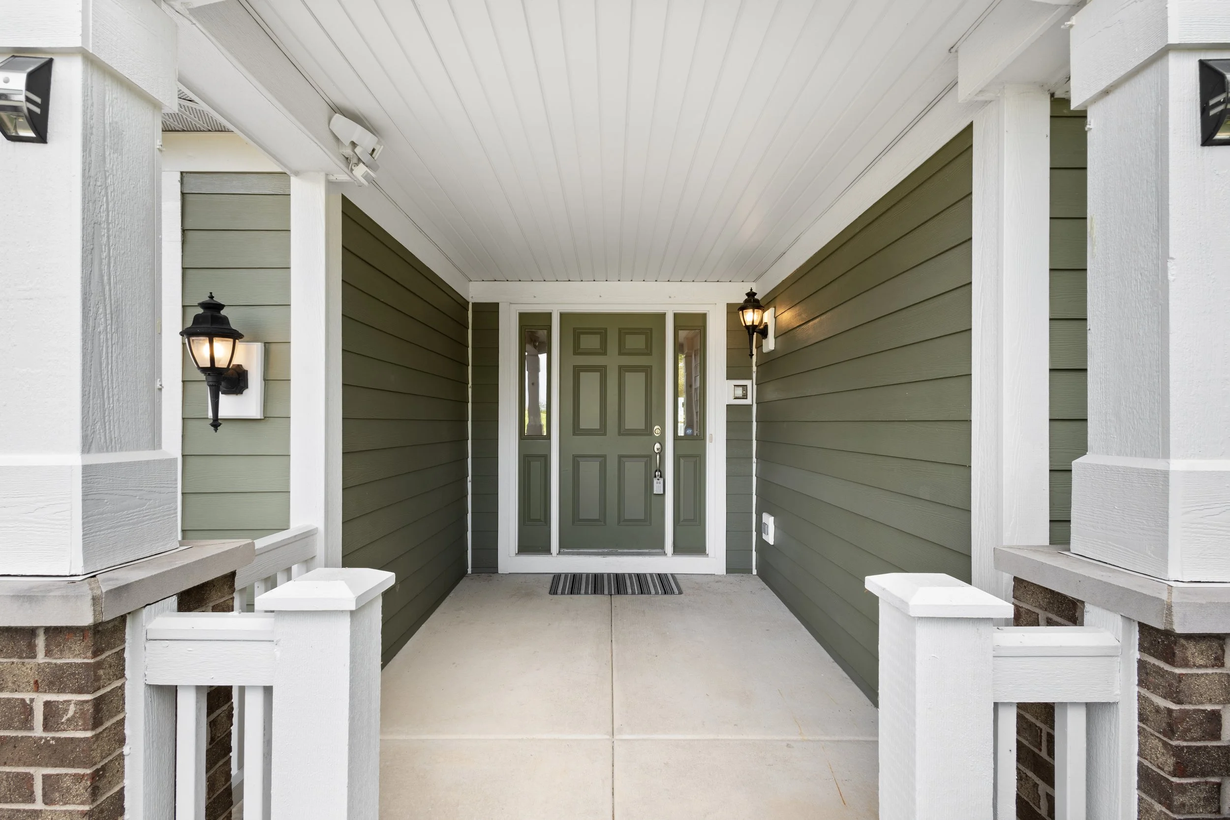 Front porch of a house with green door, brick columns, white trim, wall sconces, and a striped doormat.