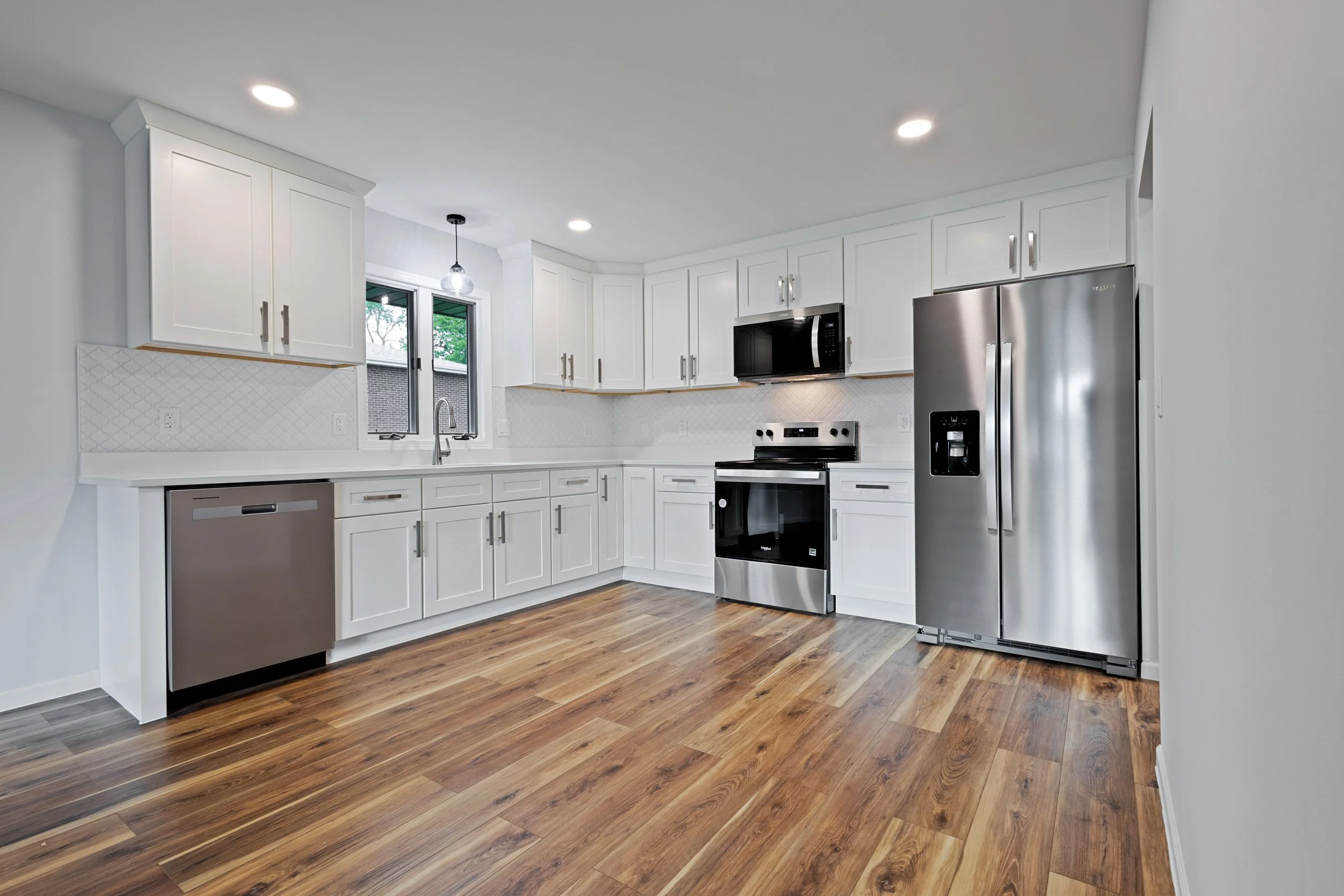 Modern kitchen with white cabinets, stainless steel appliances including a refrigerator, oven, microwave, and dishwasher, and wooden flooring.