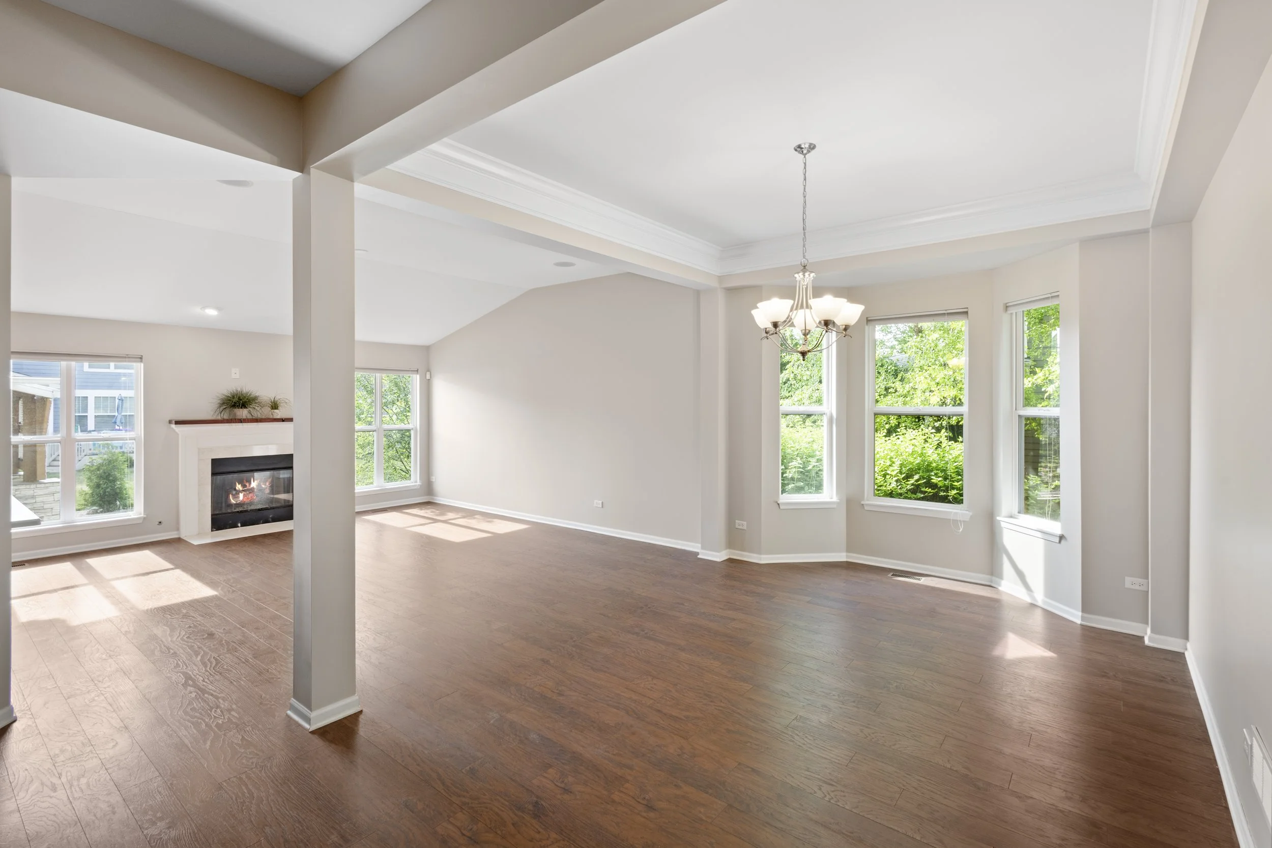 Empty living room with hardwood floors, large windows, a fireplace, and a chandelier in the dining area.