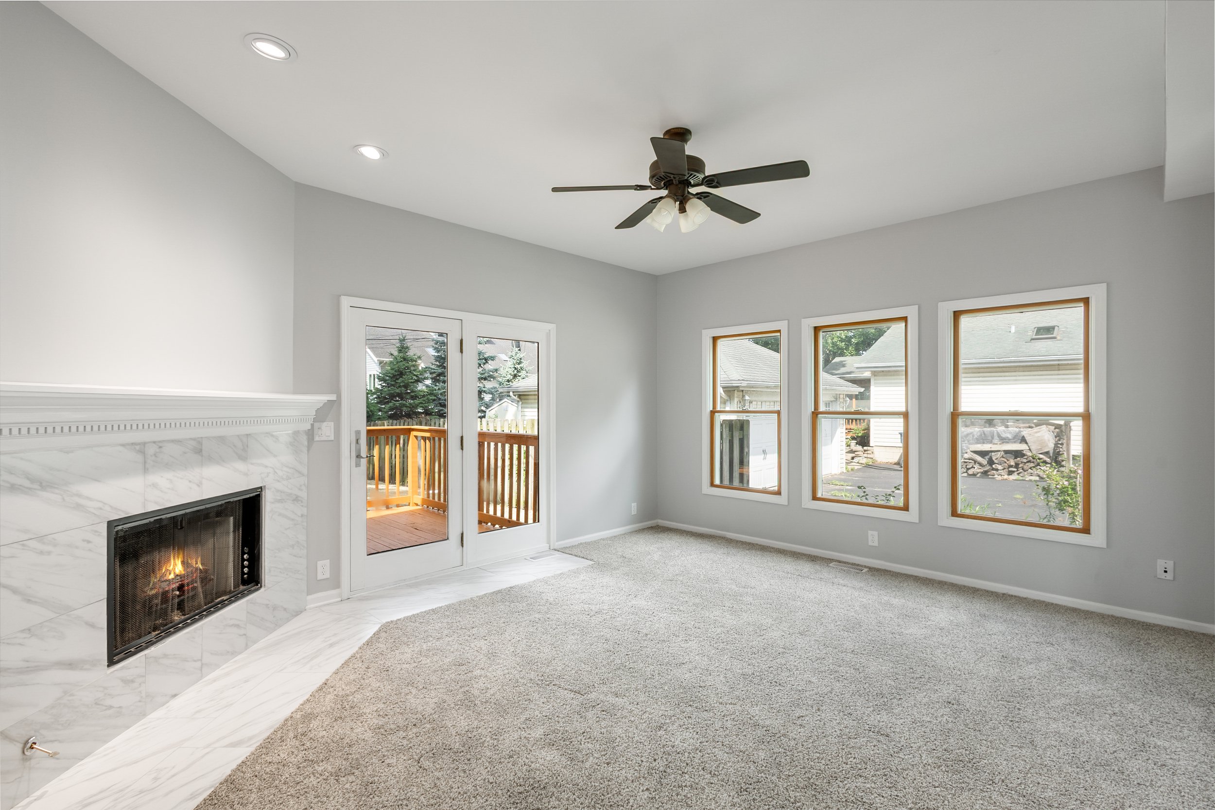 Empty living room with a fireplace, three large windows, a ceiling fan, and a sliding glass door leading to a wooden deck, with a view of neighboring houses and trees outside.