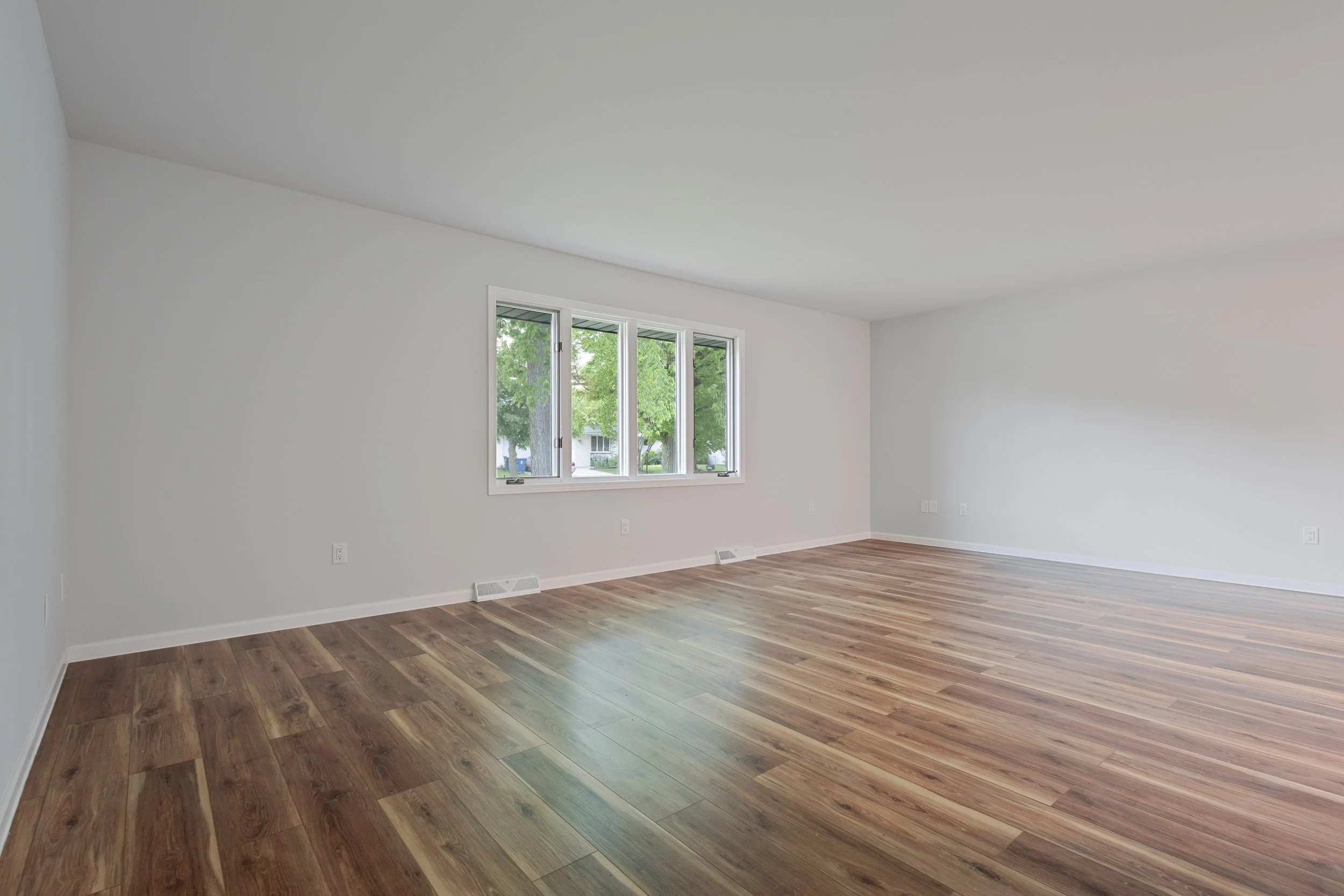 Empty living room with white walls, wooden flooring, and a window showing trees outside.