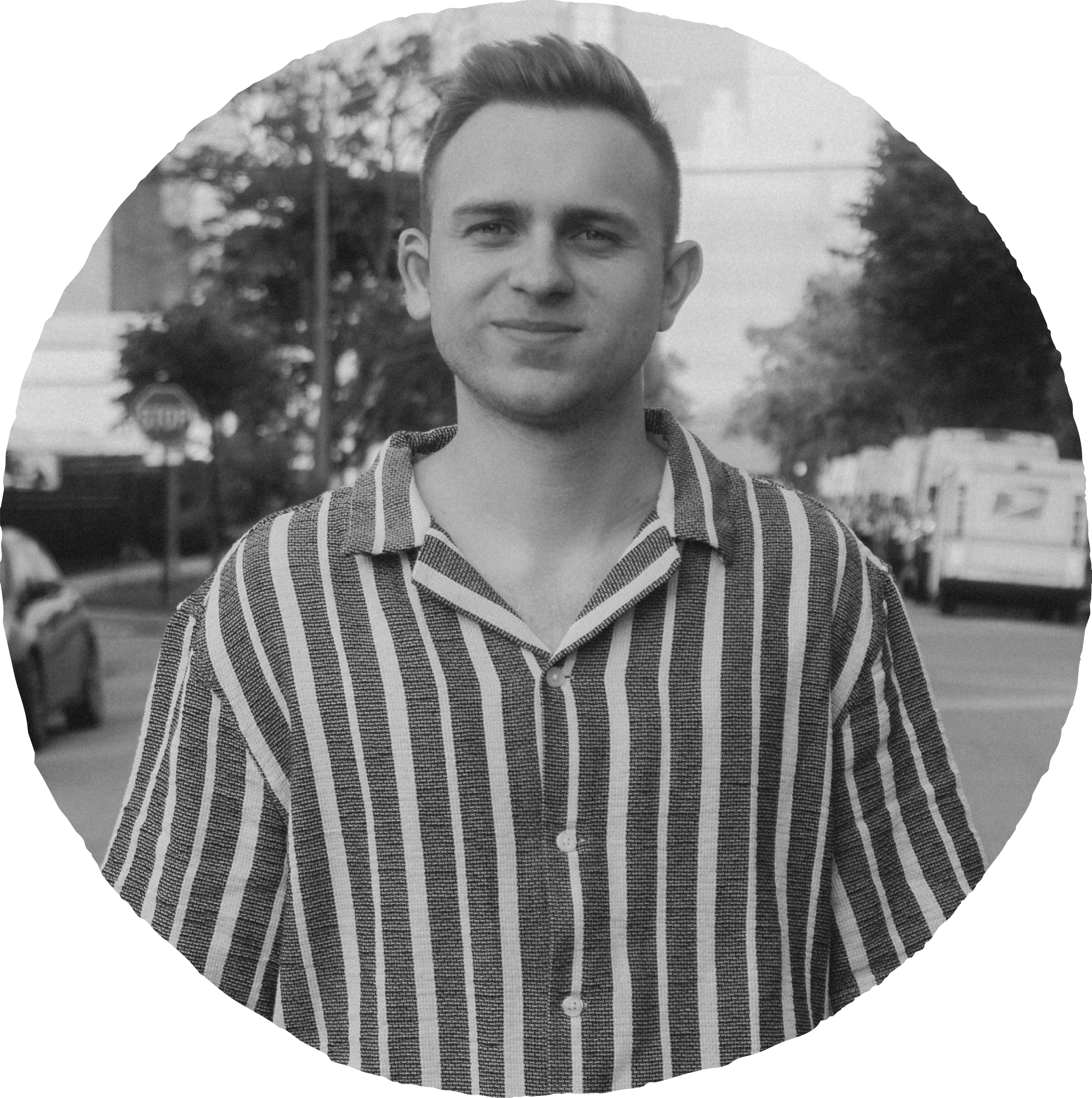 Black and white photo of a young man outdoors, wearing a striped shirt, standing on a street with trees and vehicles in the background.