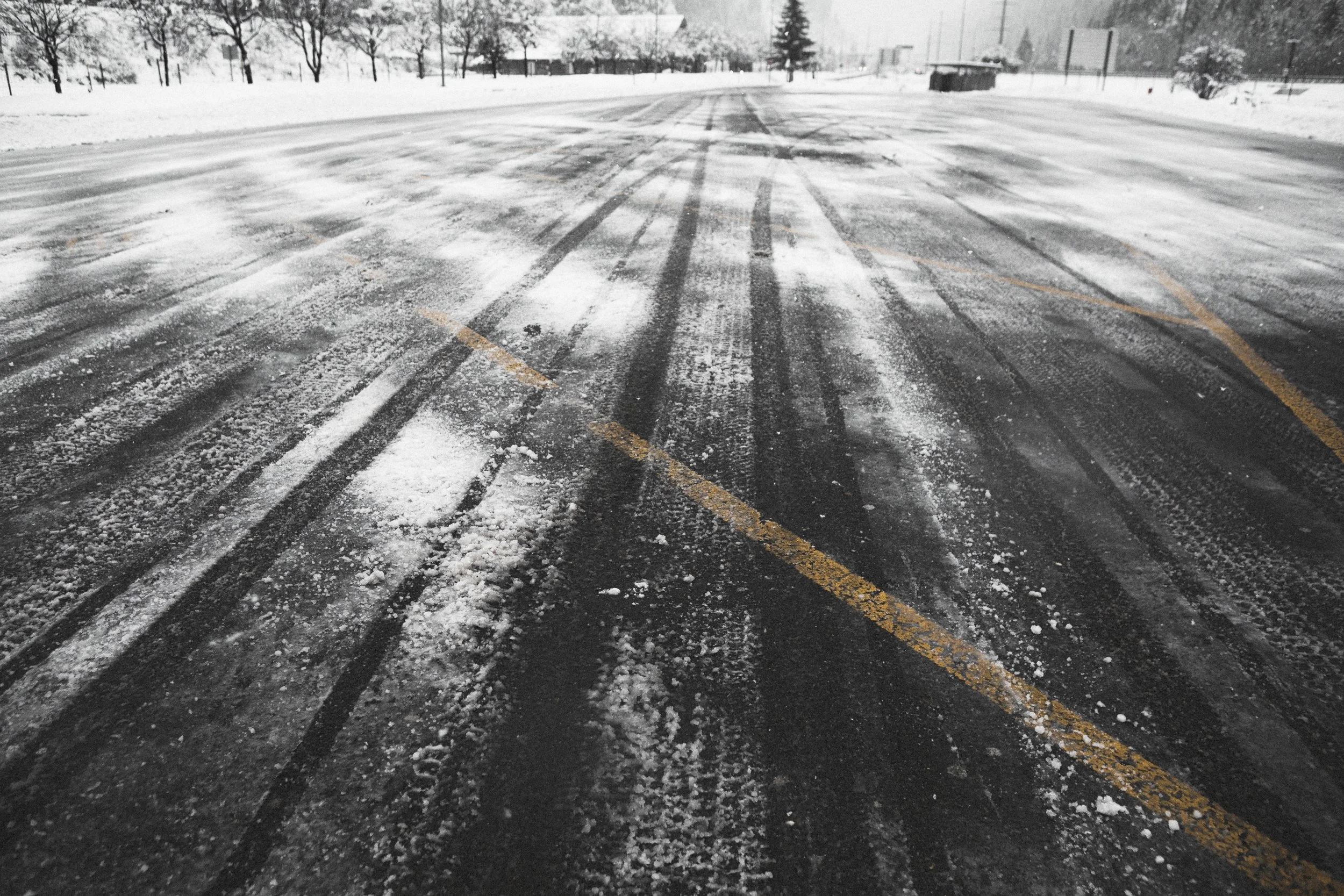 Snow-covered parking lot with tire tracks on the icy surface, surrounded by snow-covered trees and distant buildings.