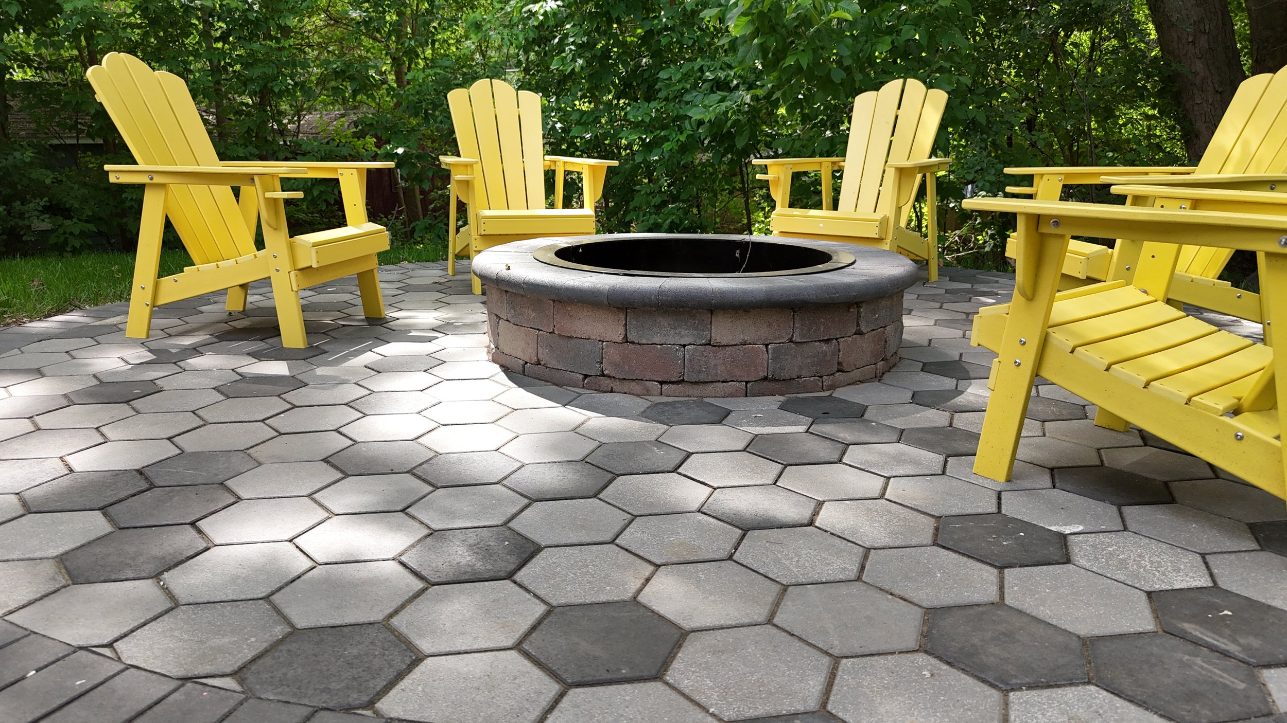 Six yellow chairs arranged around a King Maple installed brick fire pit on a hexagonal tile patio, surrounded by green trees.