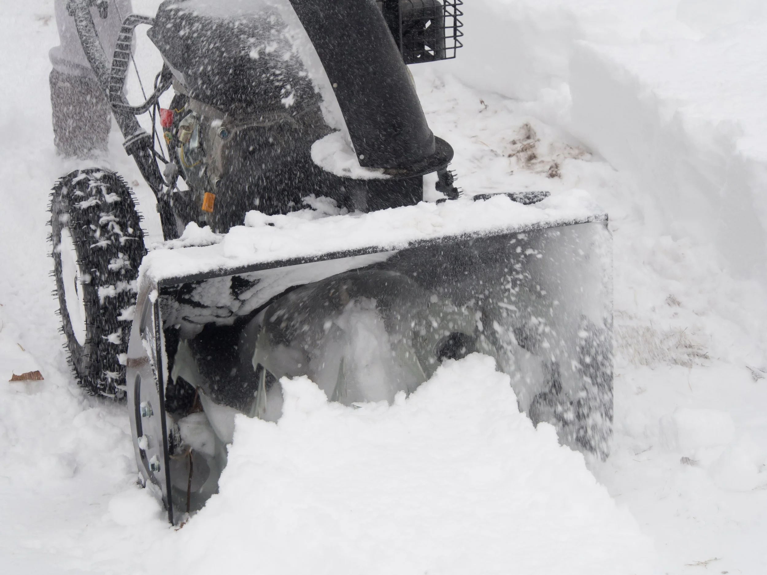 A snowblower clearing snow from a sidewalk during a snowstorm, with snow flying through the air.