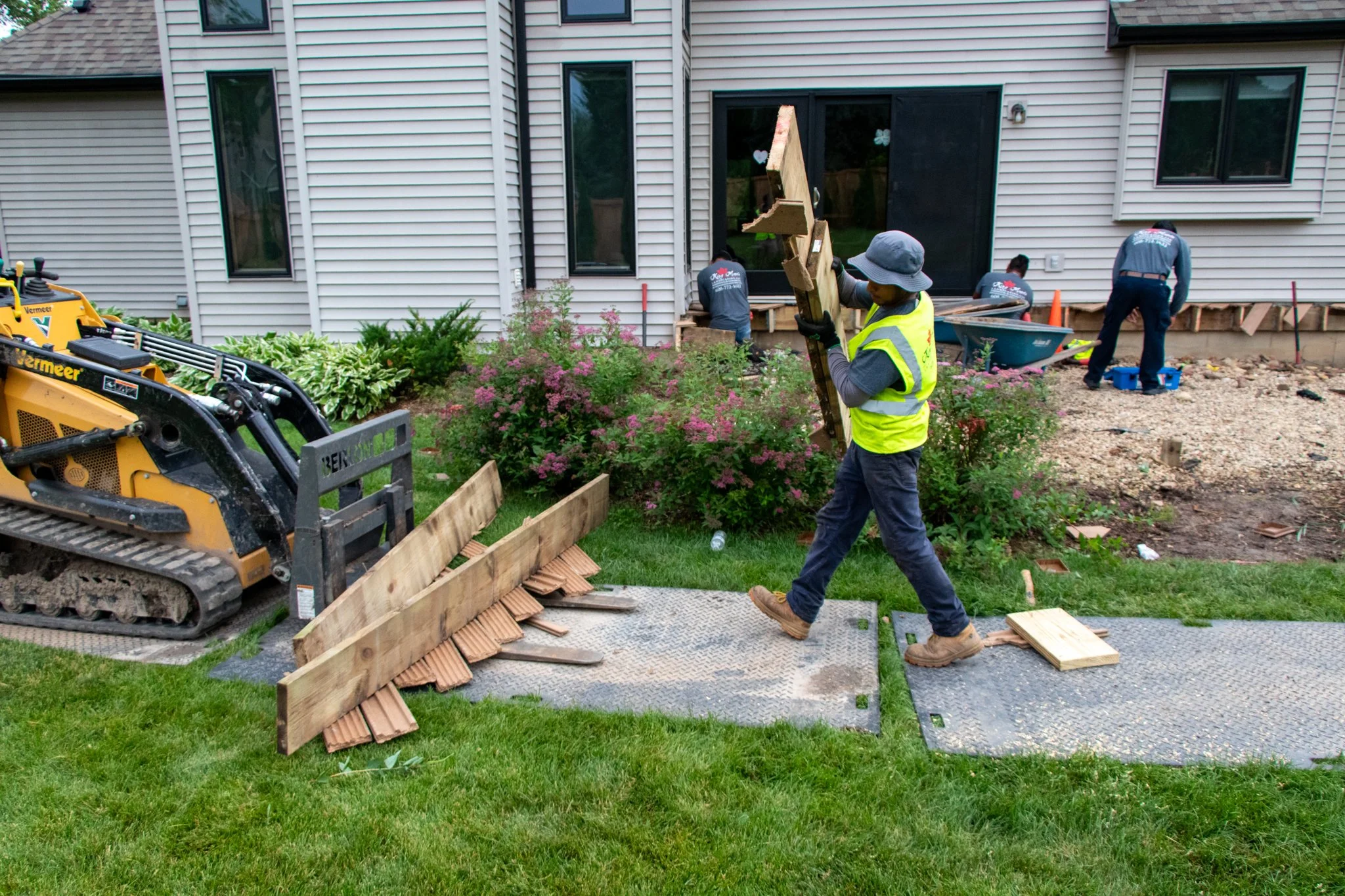 King Maple employees installing a wooden deck in the backyard of a house. One employee holds a piece of wood while others work at the deck site. There is construction equipment and materials around, including a yellow mini excavator and tools.