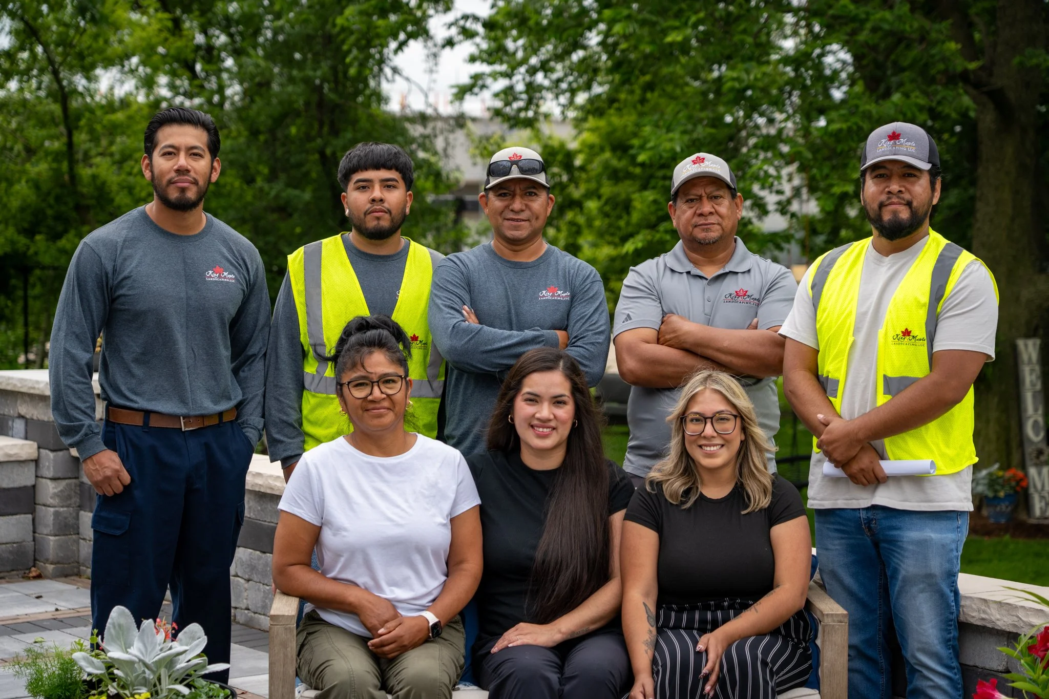 Group of diverse people posing outdoors, some wearing work vests and hats, others in casual clothes, in a park with green trees.