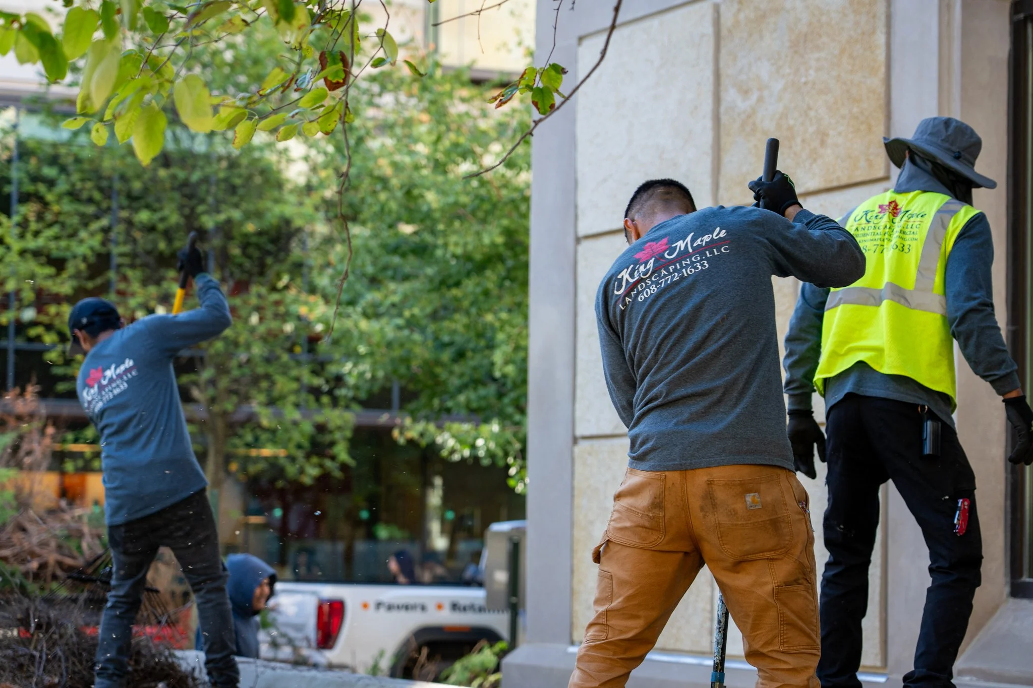 Group of workers planting or landscaping outside a beige building, some wearing gloves and hats, one wearing a high-visibility jacket, surrounded by trees and shrubs.