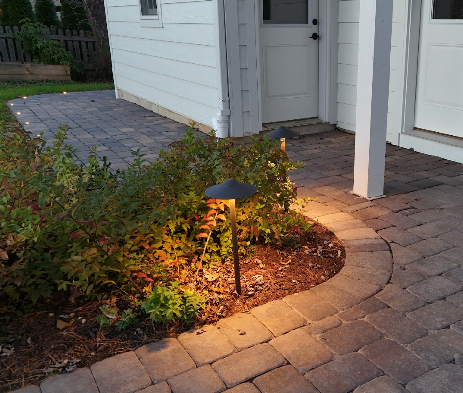 A backyard patio with stone pavers and low-voltage landscape lighting, next to a white house with a door and a small supporting column.