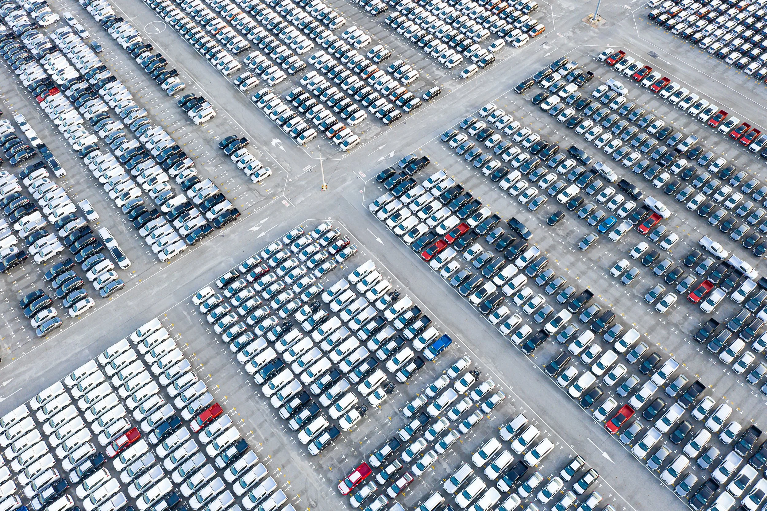 An aerial view of a large parking lot filled with rows of white, black, and some red and blue cars.
