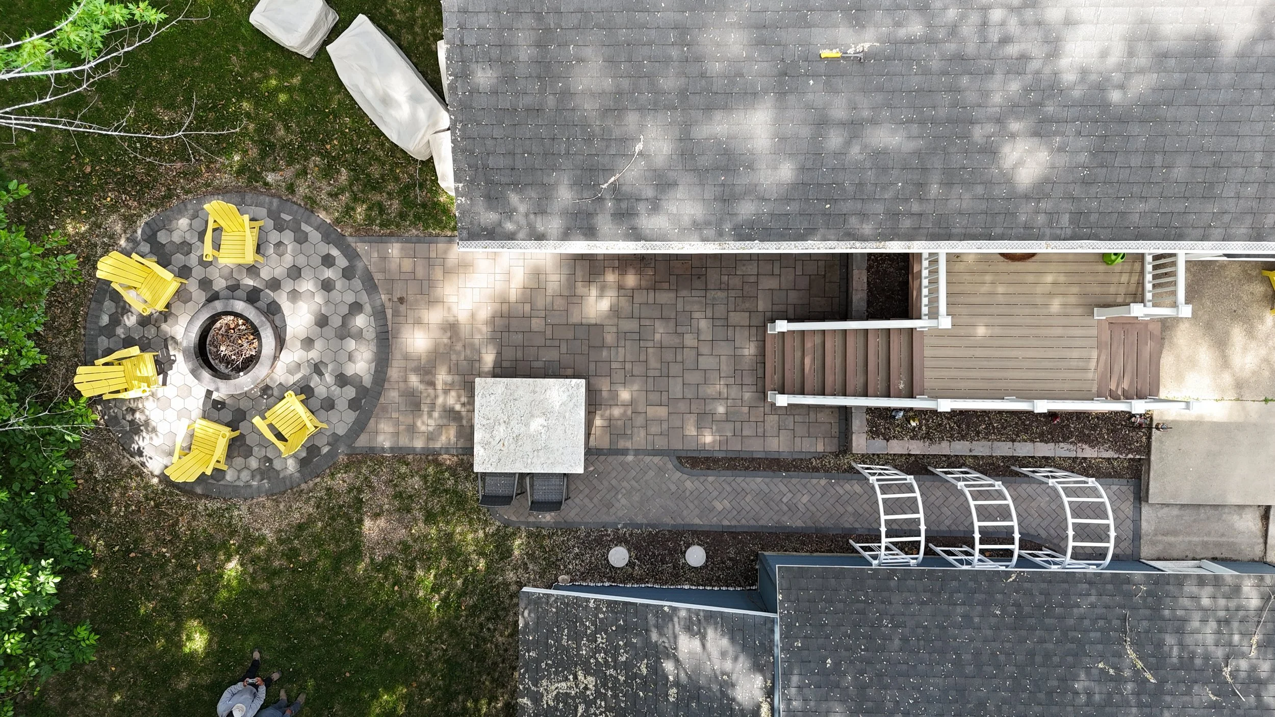 An aerial view of a backyard patio with a circular seating area containing five yellow chairs surrounding a fire pit. Adjacent to the patio is a house with a gray shingled roof and a small wooden deck with stairs. There are trees and grass in the yard, and two people are standing on the grass at the bottom of the image.