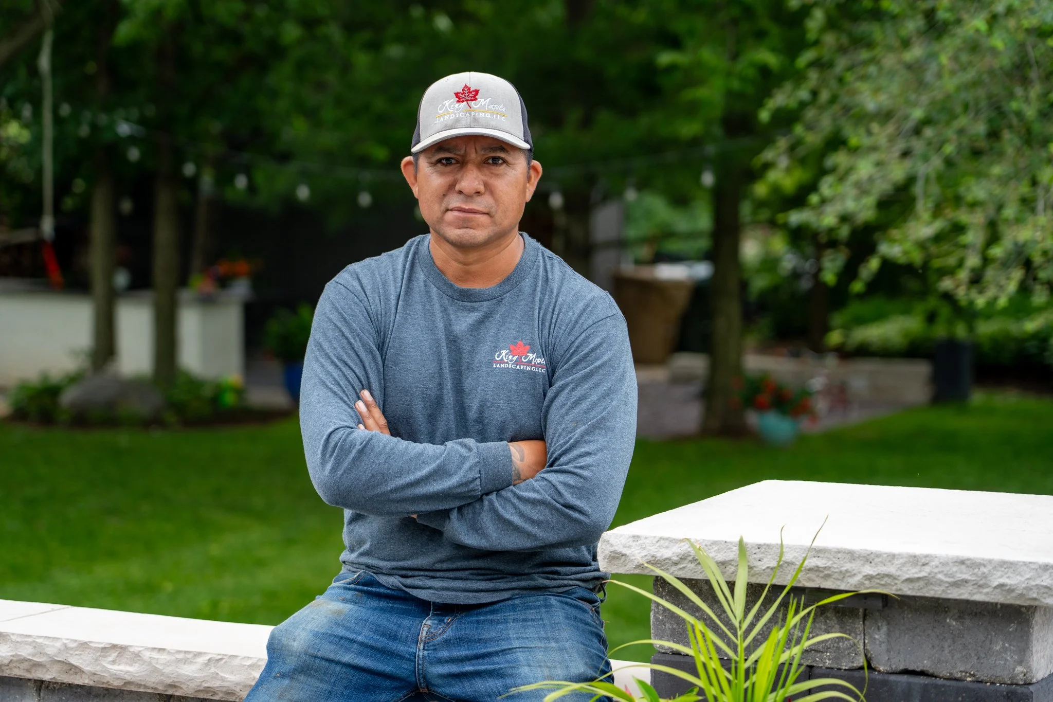 King Maple employee with crossed arms, wearing a gray and black baseball cap with a red maple leaf and logo that reads 'King Maple Landscaping LLC,'