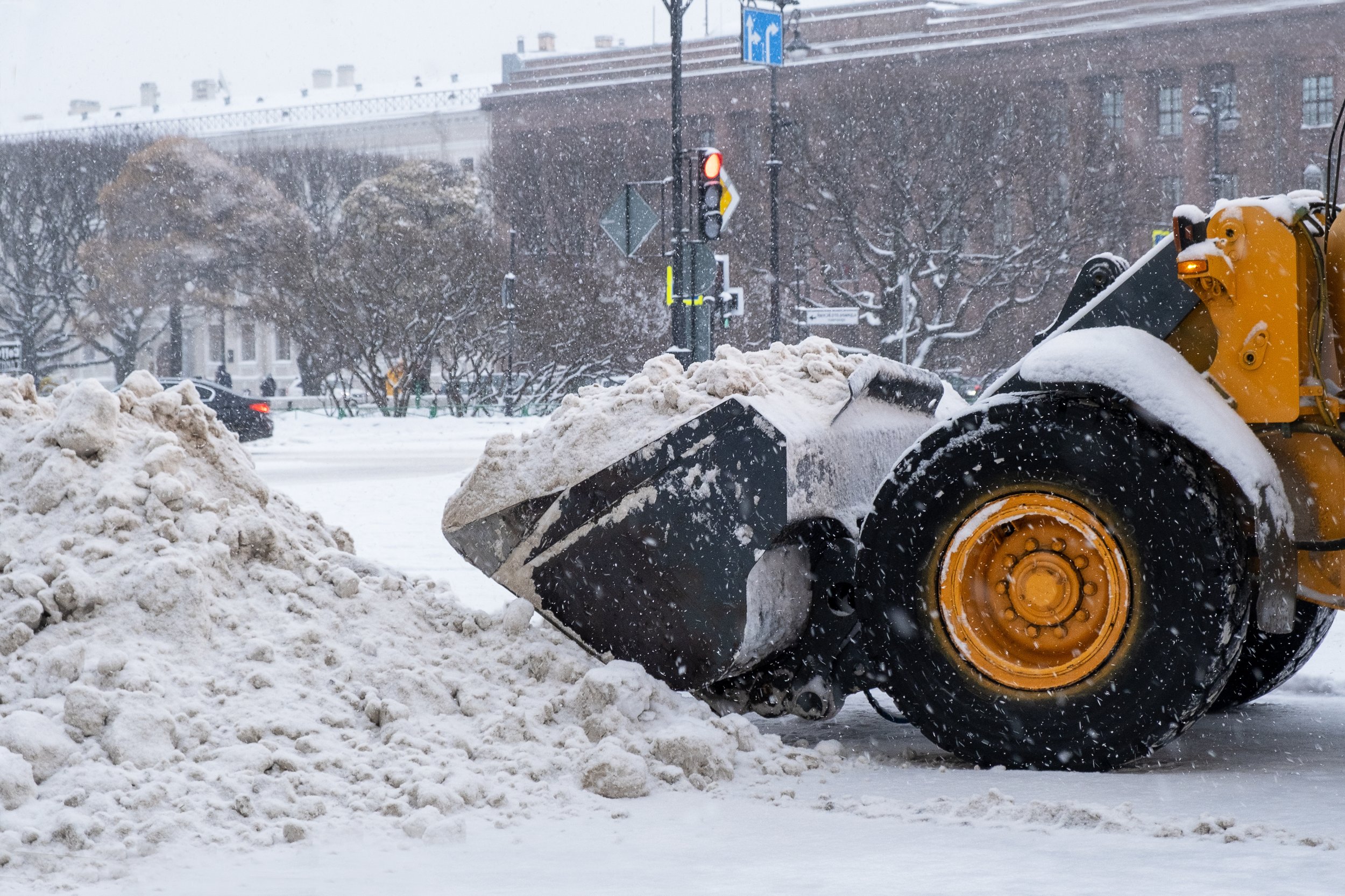 A snowplow with yellow wheels clearing snow from a city street during a snowstorm, with snow-covered trees and buildings in the background.