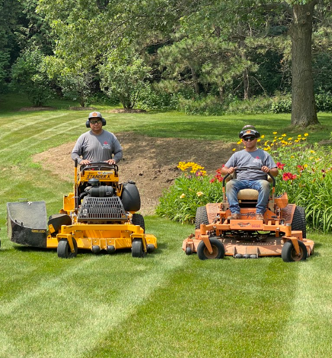 Two King Maple Employees operating large lawn mowers on a well-maintained green lawn, with trees and colorful flowers in the background.