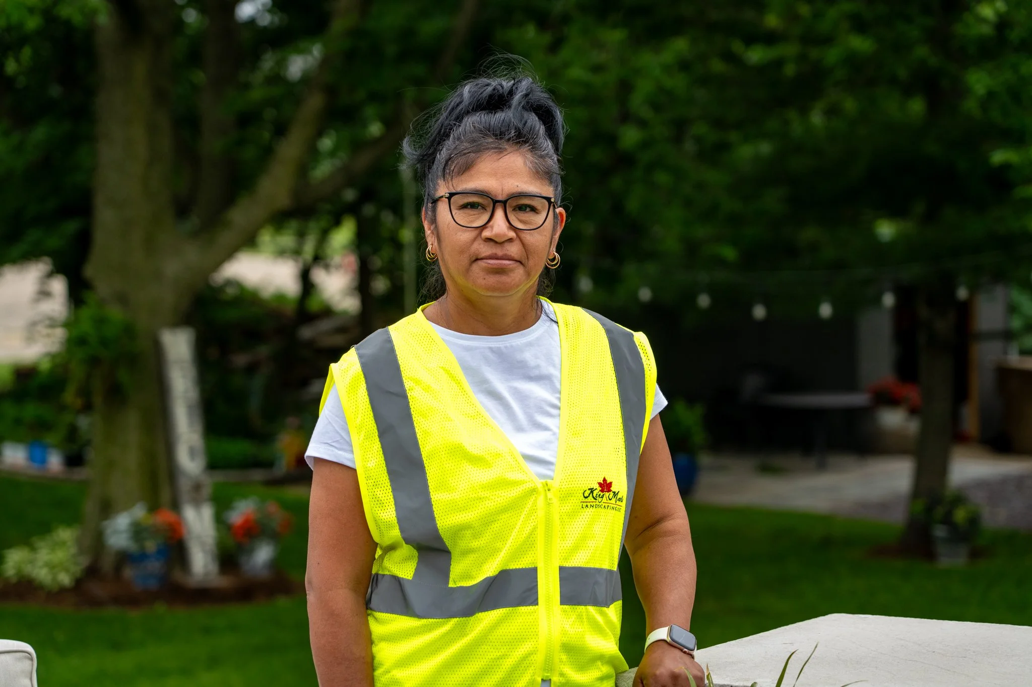 King Maple employee with glasses, dark hair tied in a high ponytail, wearing a high-visibility yellow vest over a white T-shirt, standing outdoors with trees in the background.