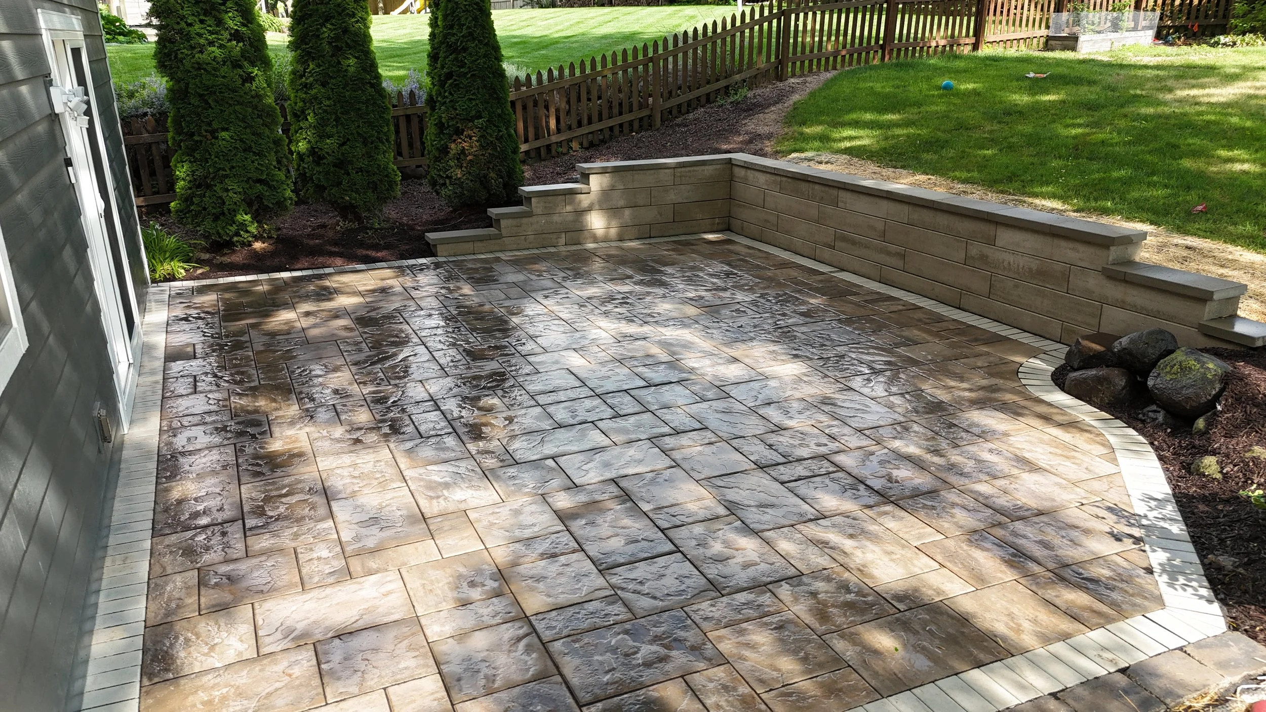 King Maple Hardscaping Project: A backyard patio with wet, stamped concrete featuring a textured pattern, bordered by paver edging, and a low stone retaining wall with steps leading to a grassy yard. There are trees and a fence in the background.