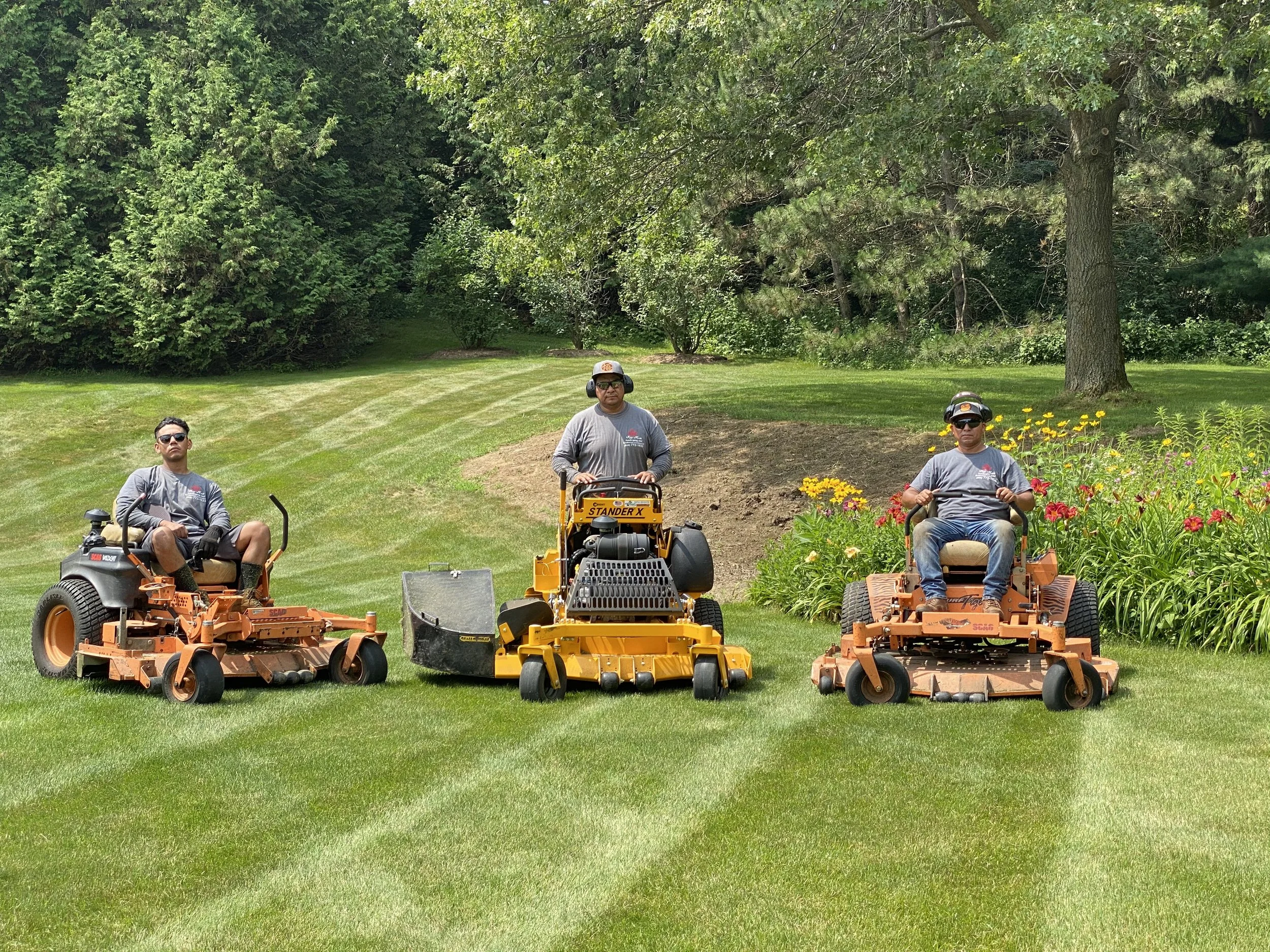 Three King Maple Employees operating lawn mowing equipment on a well-maintained grassy yard with large trees and colorful flower beds in the background.
