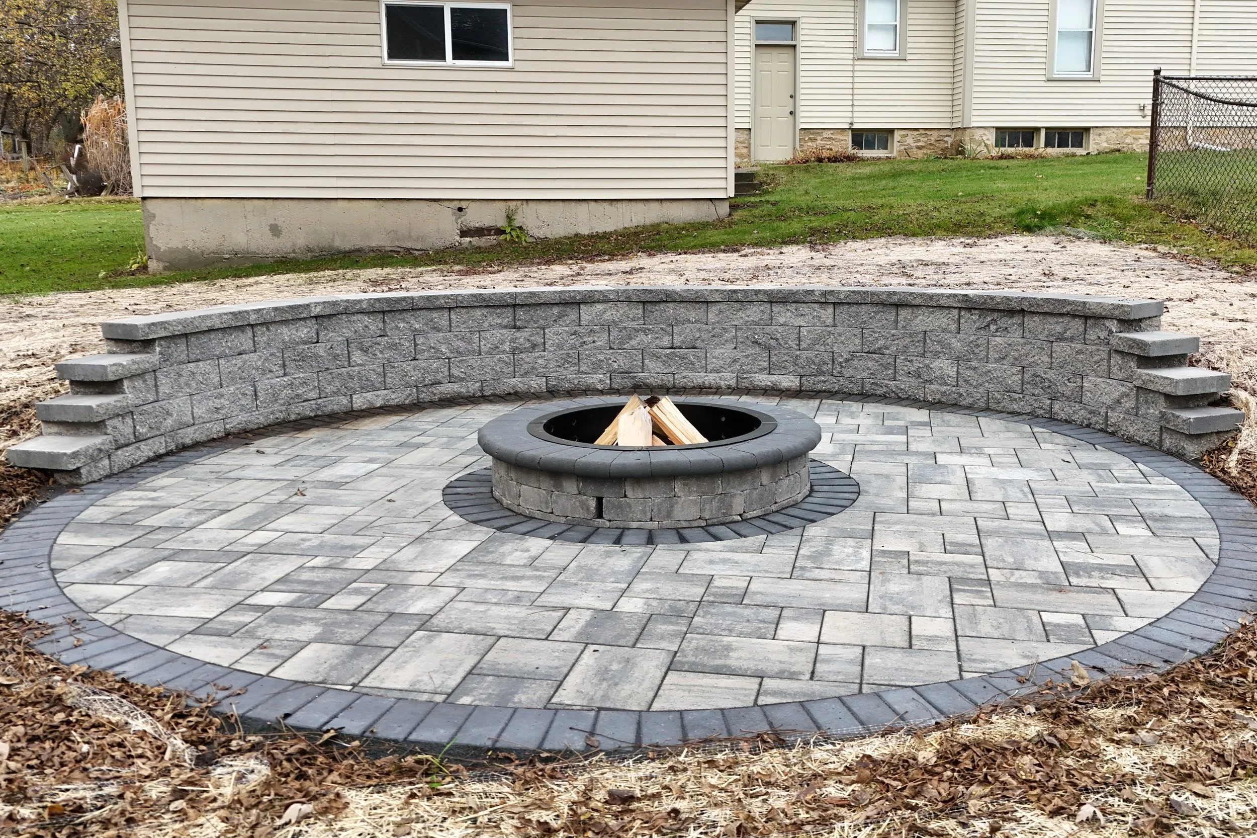 King Maple Hardscaping Job: A backyard with a new circular stone fire pit surrounded by gray brick pavers and a curved retaining wall, with a house in the background.