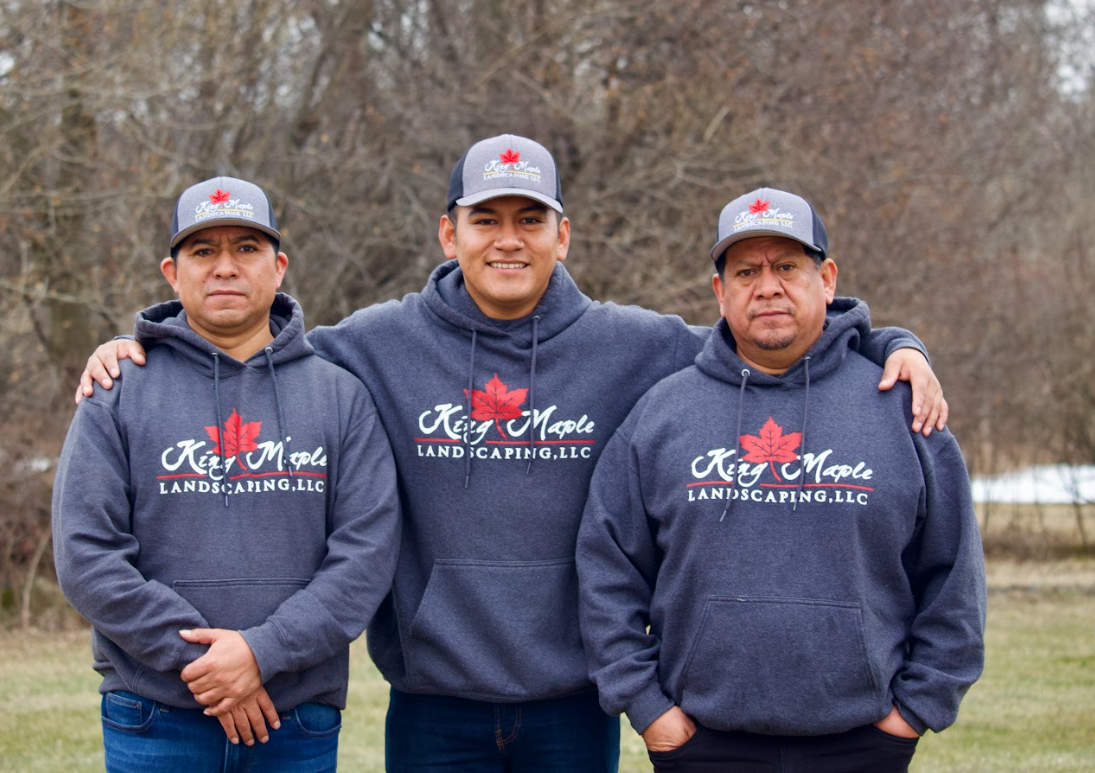 Three King Maple employees wearing matching hoodies and caps with the logo 'King Maple Landscaping, LLC' standing outdoors with trees in the background.
