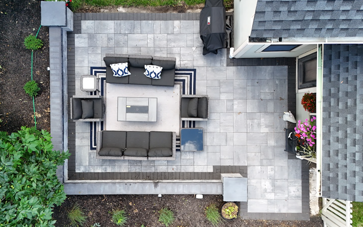 Top-down view of King Maple Hardscaping Job: an outdoor patio with gray tile flooring, seating area with grey sofas and armchairs, a coffee table, potted plants, and a grill covered with a black cover.