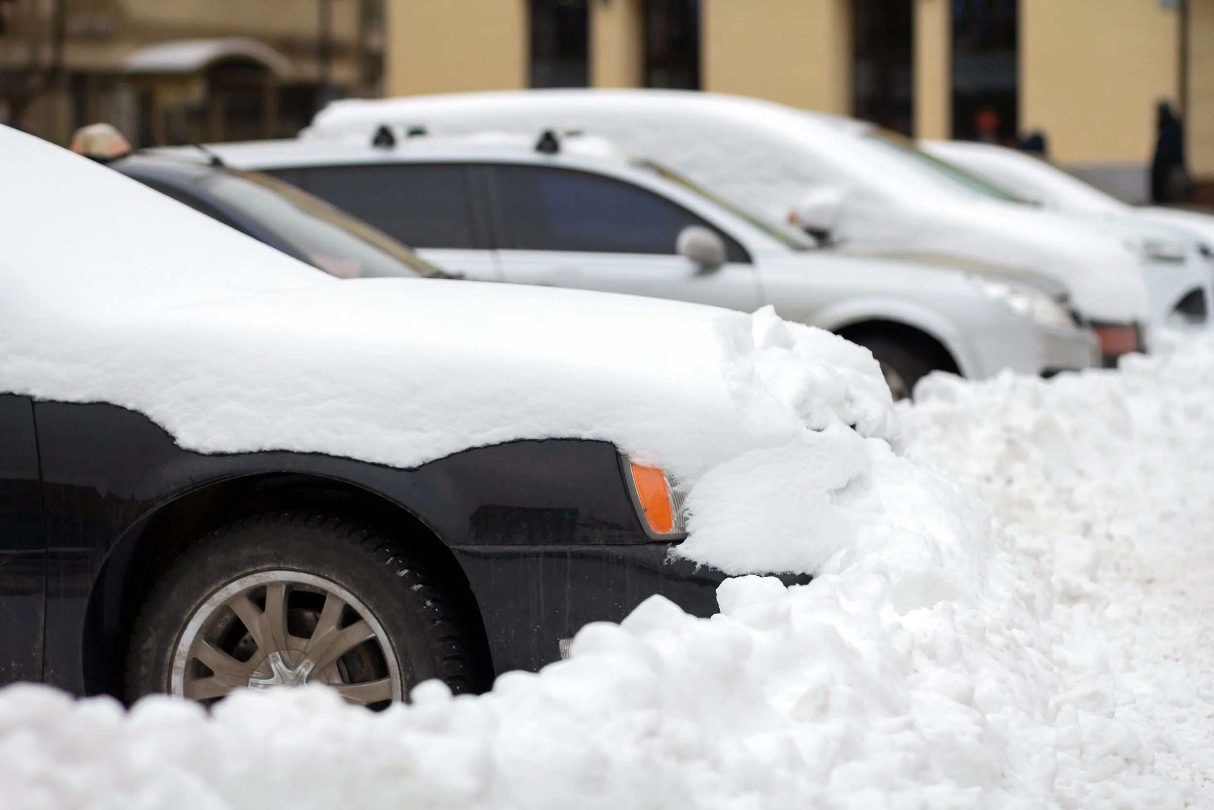 Snow-covered black car parked on the street with others in the background, and footprints in the snow.